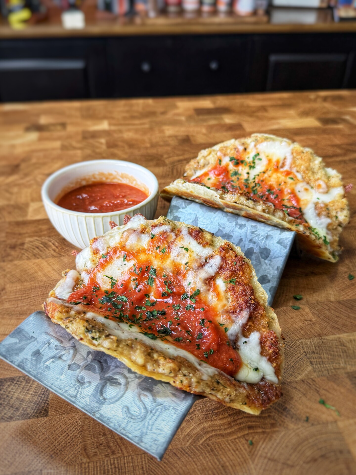 Two taco-shaped dishes made with a baked cheese shell, filled with tomato sauce and melted cheese, are displayed on a wooden counter. A small bowl of marinara sauce sits nearby for dipping.