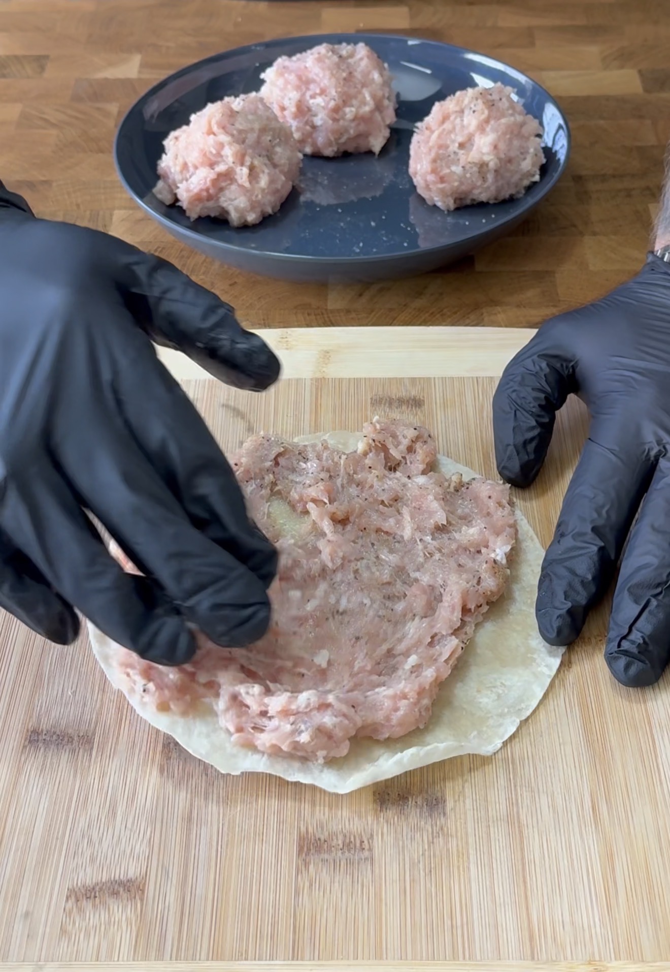 Two hands in black gloves spread raw ground meat onto a round wrapper on a wooden cutting board. In the background, a plate holds several scoops of raw meat mixture.