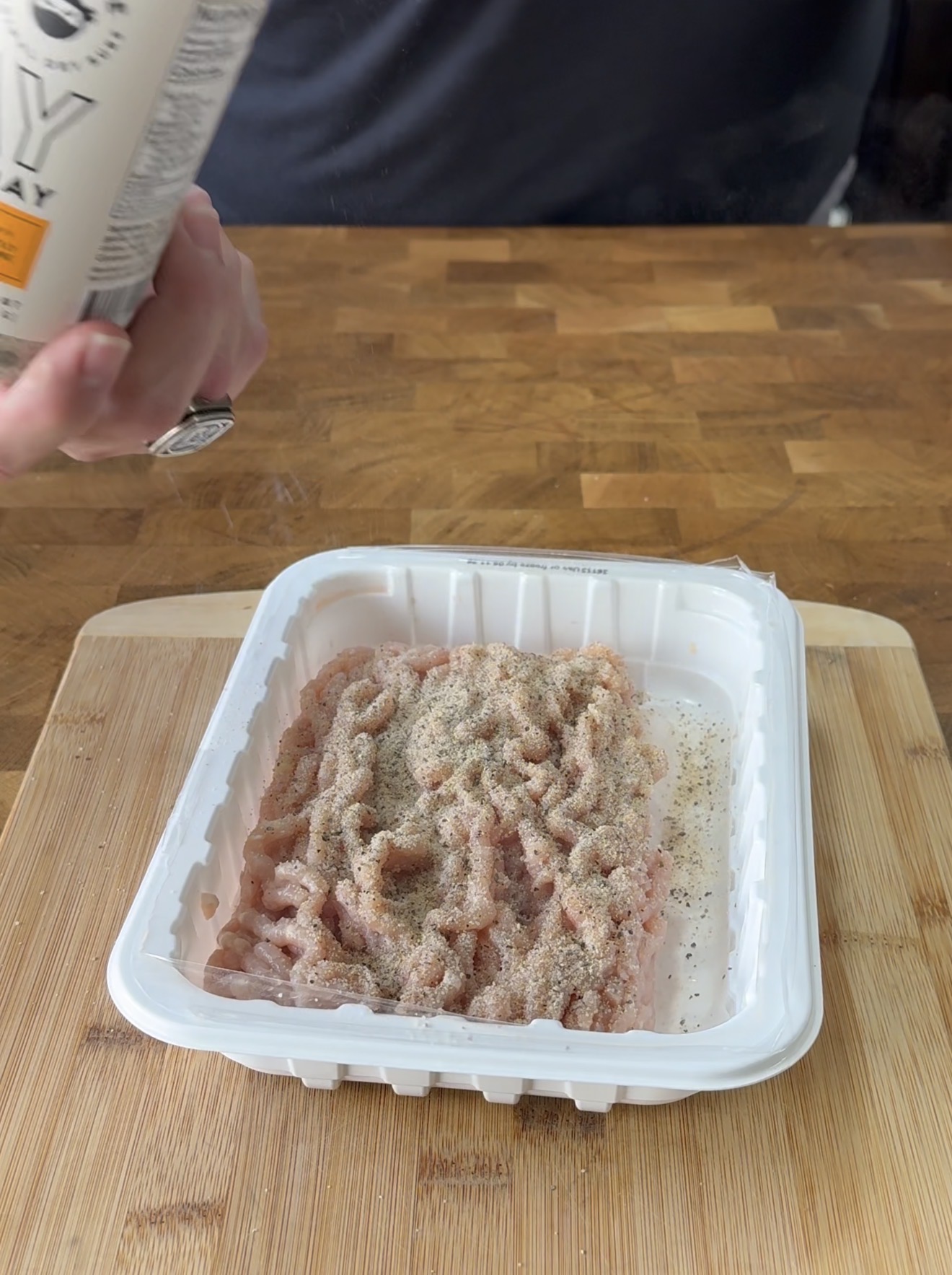 A person sprinkles seasoning onto raw ground meat in a white plastic tray placed on a wooden cutting board.