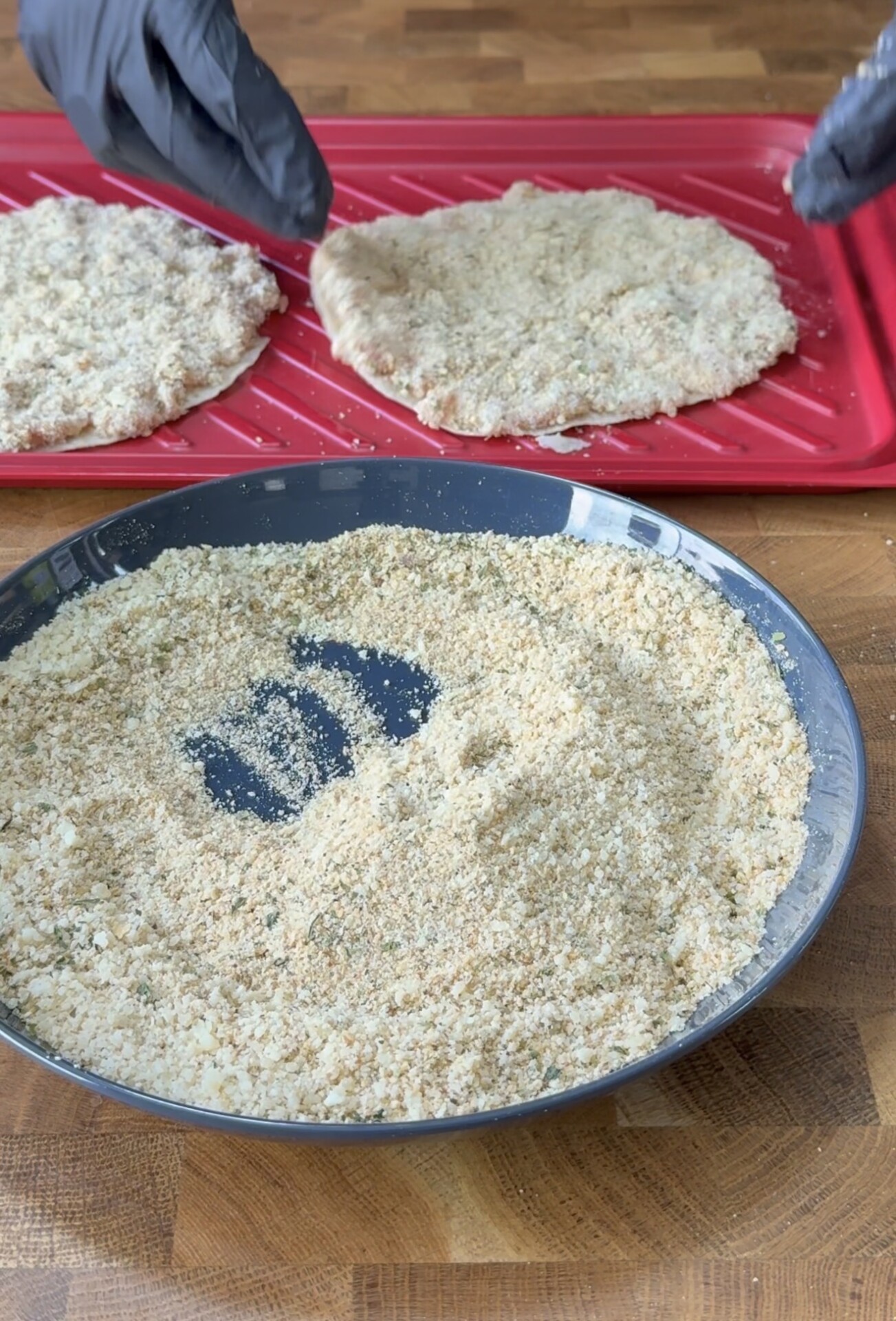 A bowl of breadcrumbs sits on a wooden counter with two breaded cutlets on a red tray in the background. A person wearing black gloves is working with the cutlets.