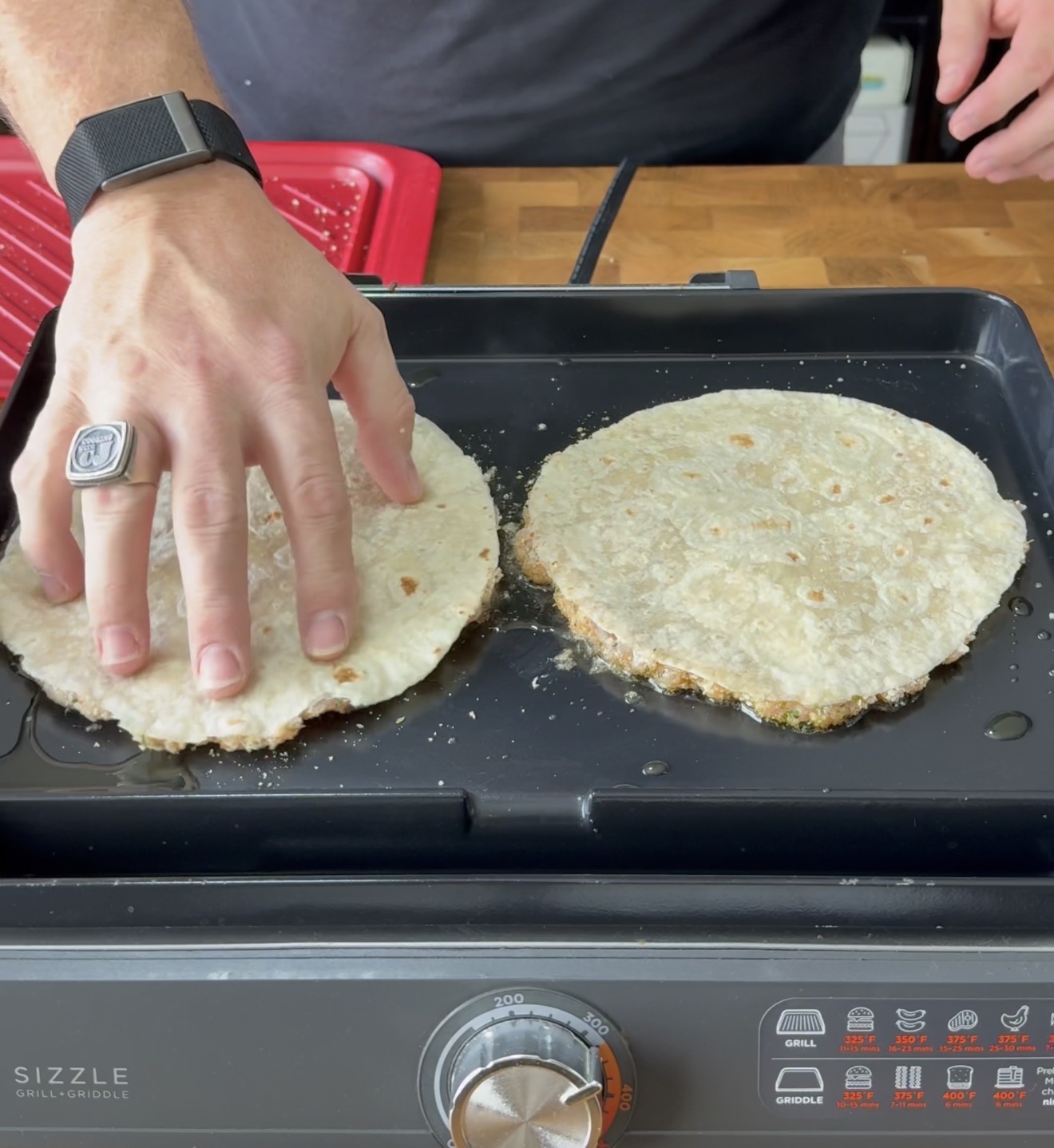 A person wearing a black watch and a ring presses a tortilla on a griddle, cooking two tortillas side by side. The griddle is on a countertop with a red tray in the background.