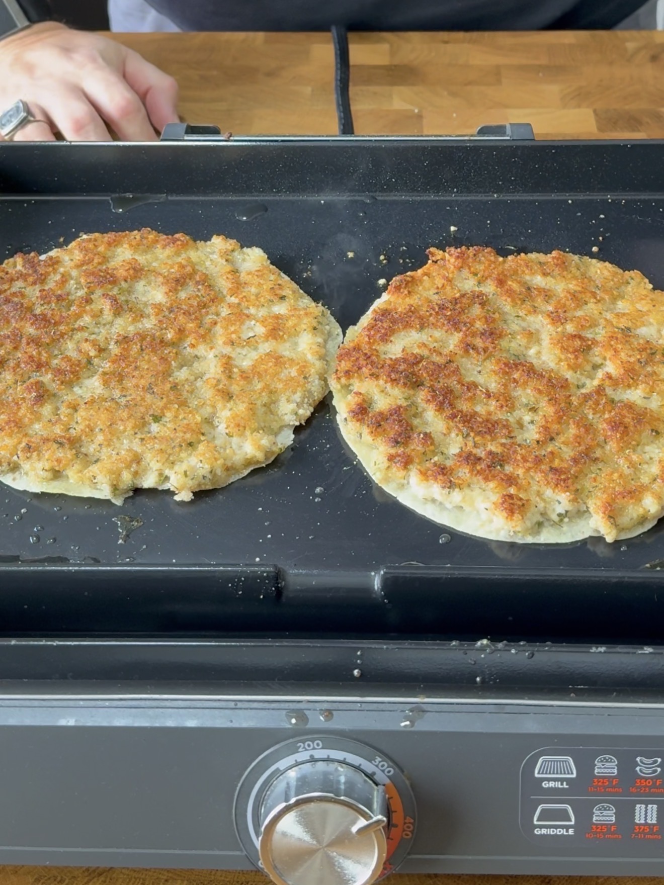 Two large, golden-brown patties are cooking on a griddle, with a person’s hands visible at the top of the image. The griddle sits on a wooden countertop.