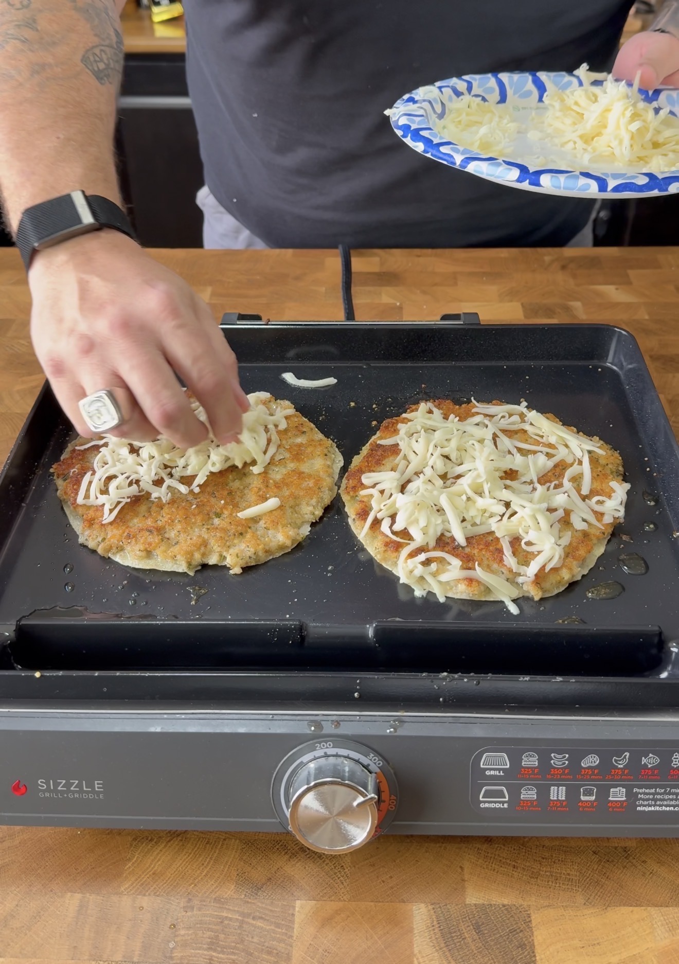 A person sprinkles shredded cheese onto two breaded patties cooking on an electric griddle. The persons arm is visible, wearing a watch and a ring, with a paper plate holding more cheese in the other hand.