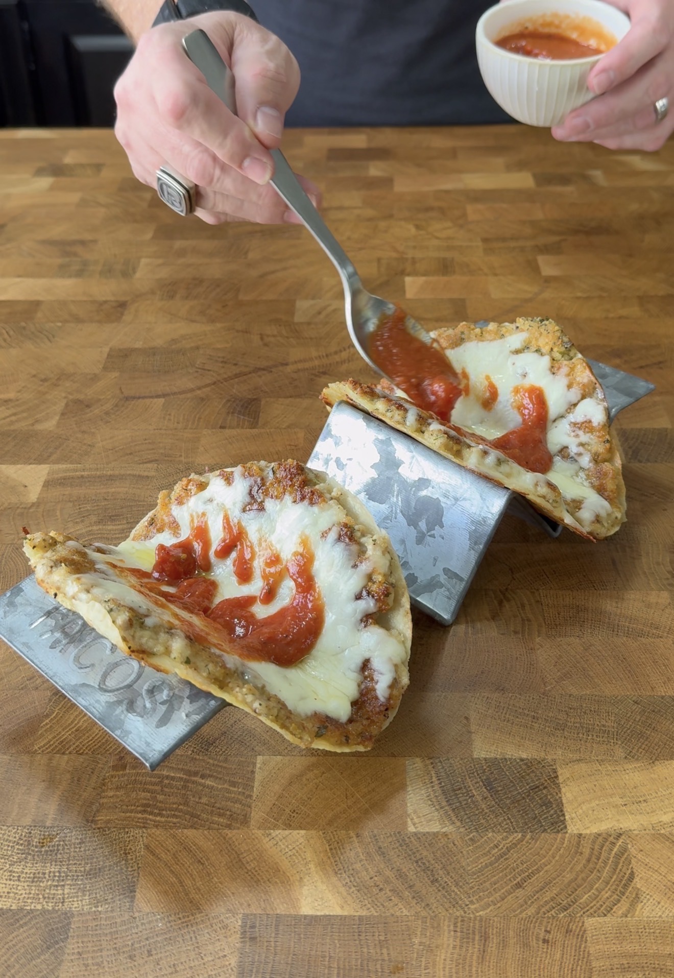 A person spoons tomato sauce onto two pizza slices folded like tacos, each held upright in a metal taco holder on a wooden countertop.