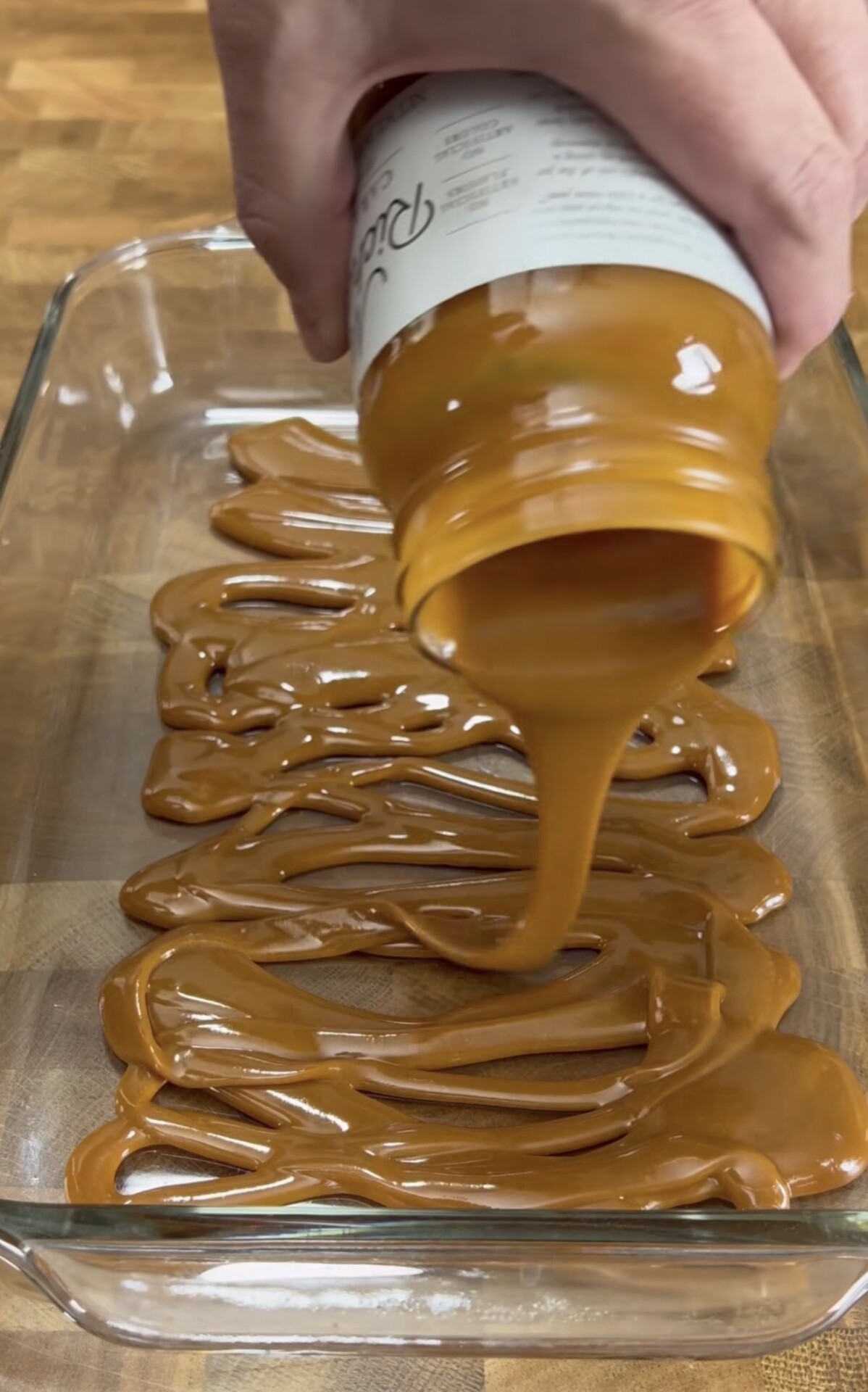 A hand is pouring thick caramel sauce from a jar into a glass baking dish, spreading it in swirls over the bottom of the dish on a wooden surface.