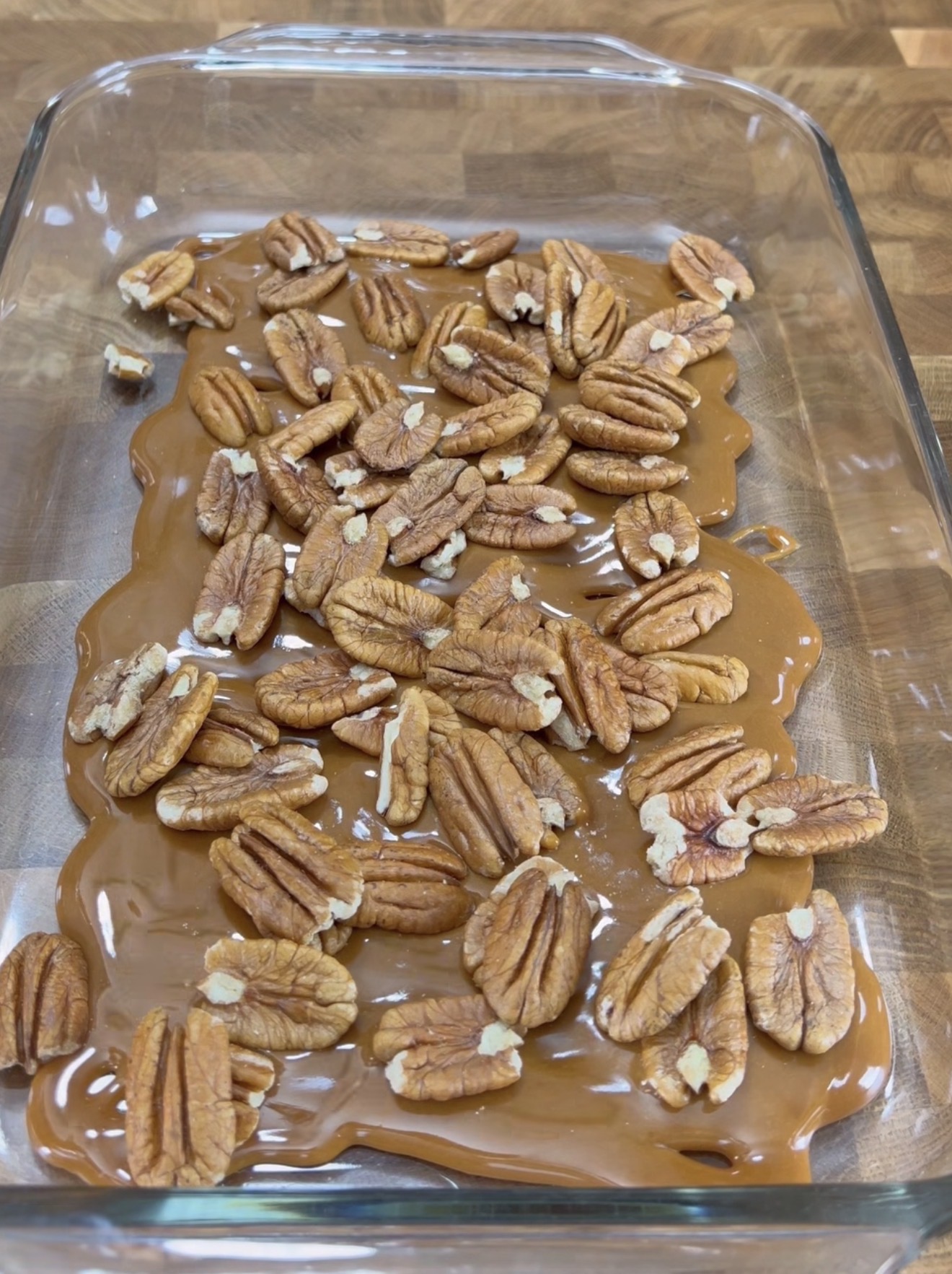A glass baking dish filled with a layer of melted caramel topped with whole pecan halves, sitting on a wooden surface.