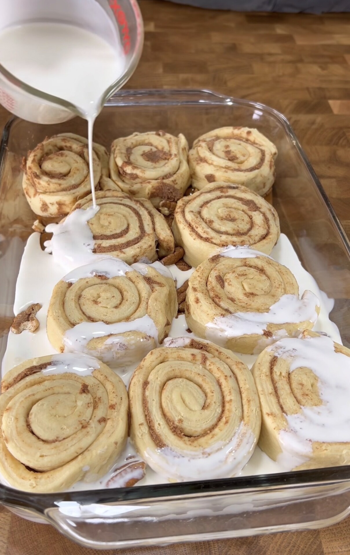 A glass baking dish filled with unbaked cinnamon rolls sits on a wooden countertop. Milk is being poured over the rolls, partially covering them before baking.