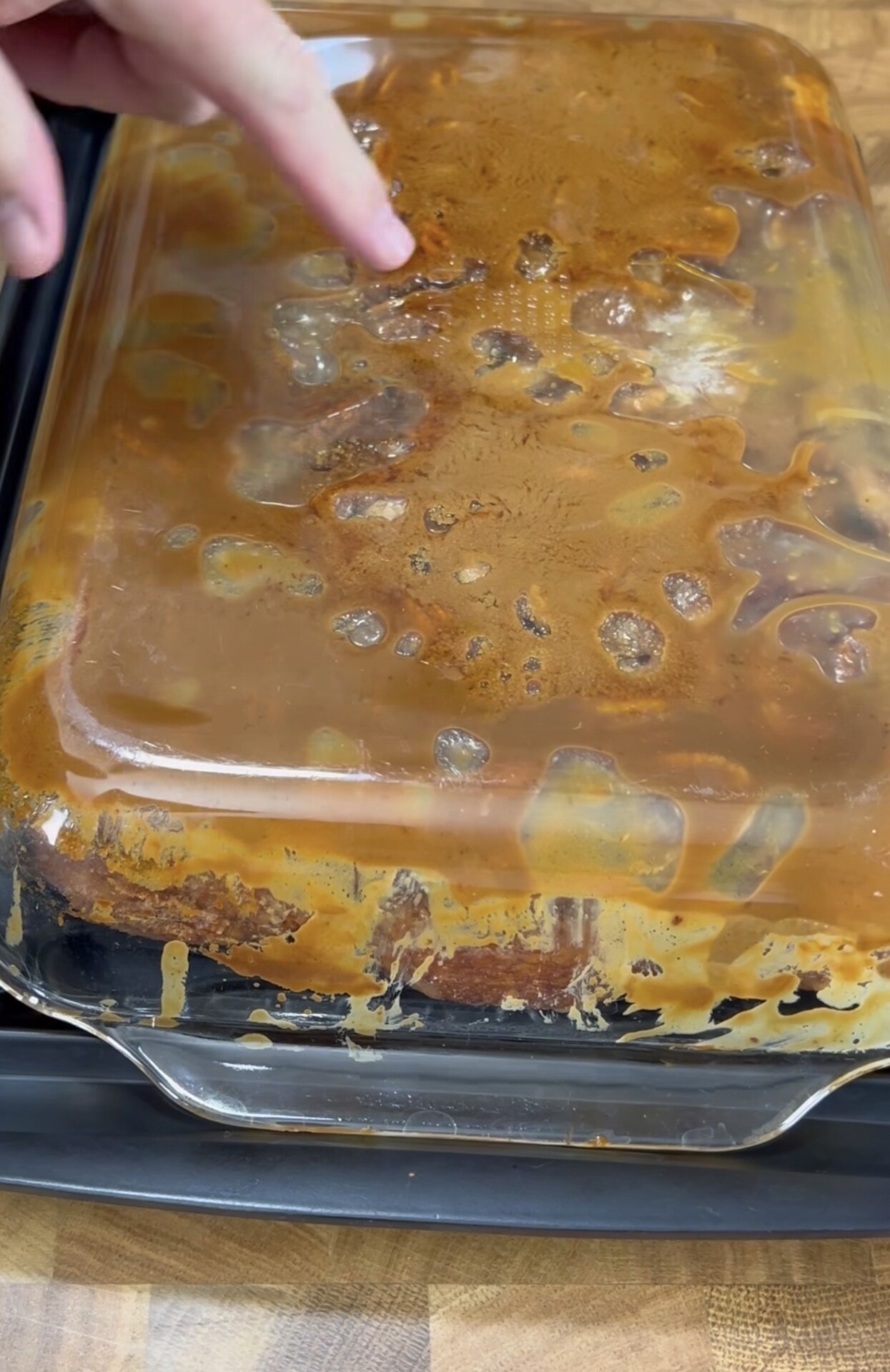A finger points at a glass baking dish covered with caramelized residue and bubbles, with streaks of sauce visible on the sides. The dish is set on a black tray atop a wooden surface.