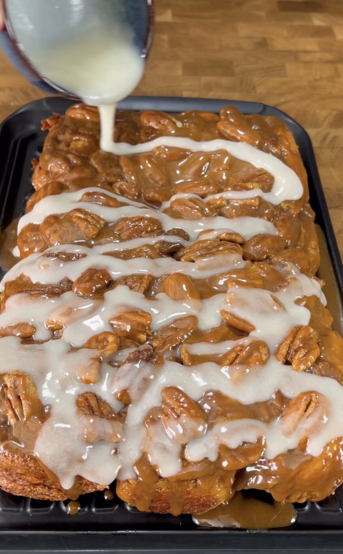 A tray of sticky buns topped with pecans and caramel, being drizzled with white icing from a bowl. The buns are set on a black baking tray atop a wooden countertop.