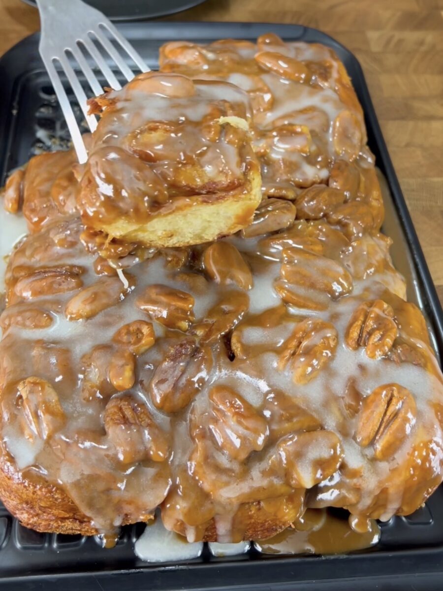 A tray of sticky pecan cinnamon rolls topped with caramel glaze and whole pecans. A spatula lifts one roll, showing the gooey, glazed texture and layers inside.