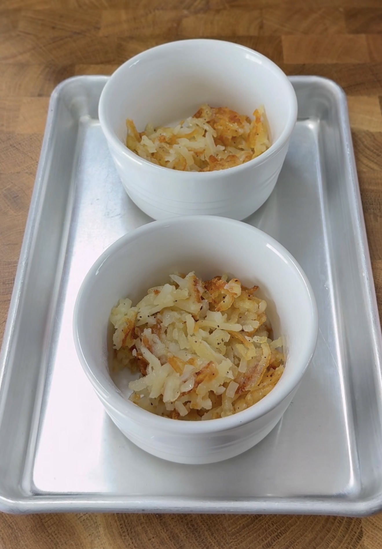 Two white ramekins filled with crispy, golden-brown hash browns are placed on a silver baking tray atop a wooden surface.