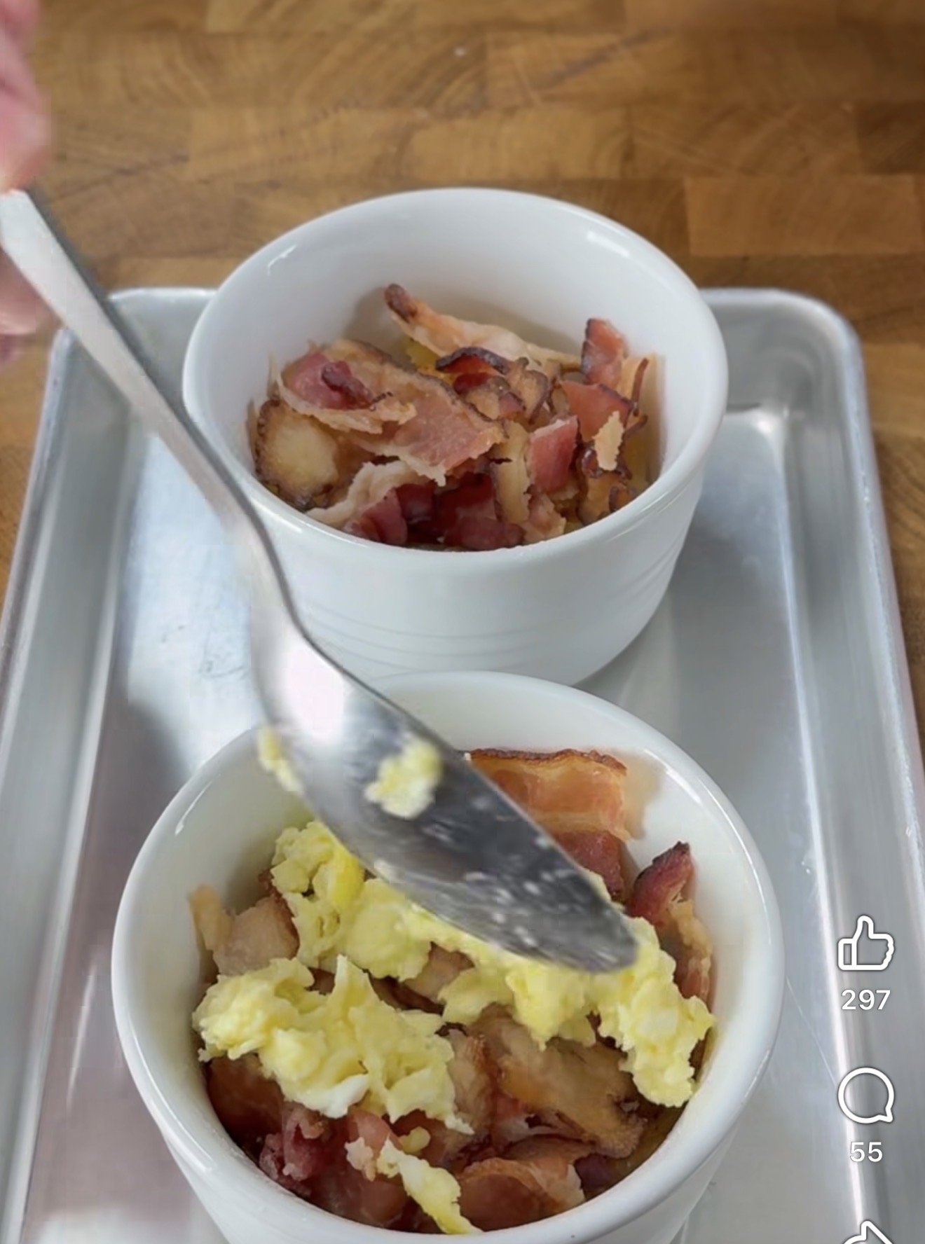 A hand uses a spoon to add scrambled eggs to a white ramekin filled with cooked bacon pieces, next to another ramekin with just bacon, both placed on a metal tray on a wooden surface.