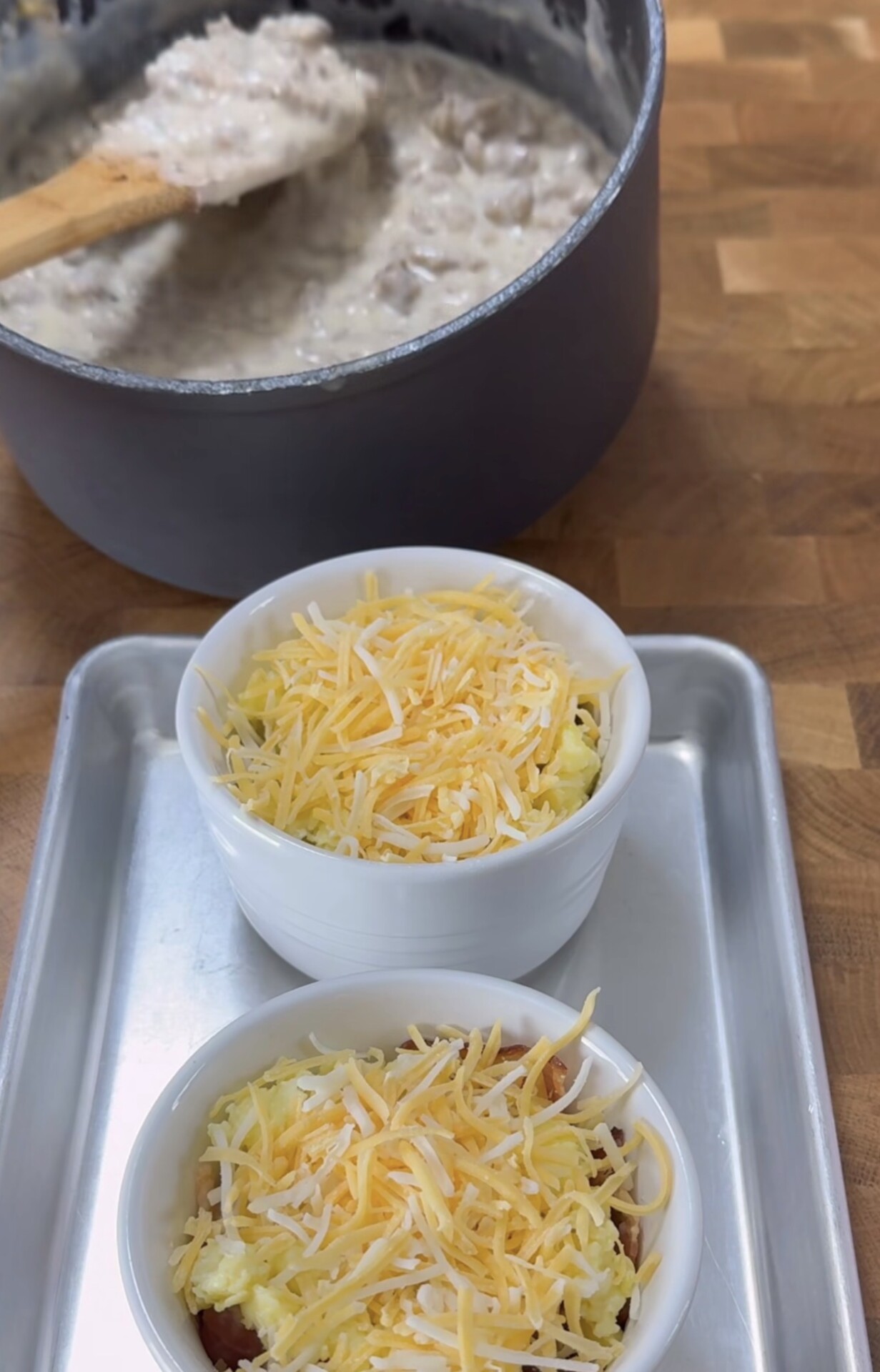 Two white ramekins filled with scrambled eggs and shredded cheese sit on a metal tray, while a pot of creamy sausage gravy with a wooden spoon is visible in the background on a wooden countertop.