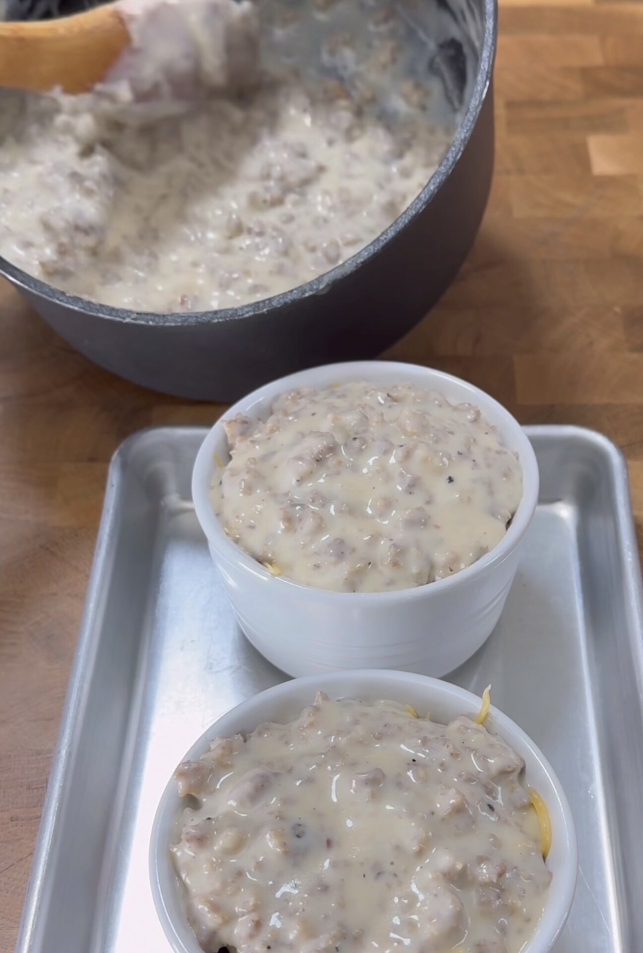 Two white ramekins filled with a creamy sausage mixture sit on a metal tray. Behind them is a pot containing more of the same mixture, with a wooden spoon resting inside.