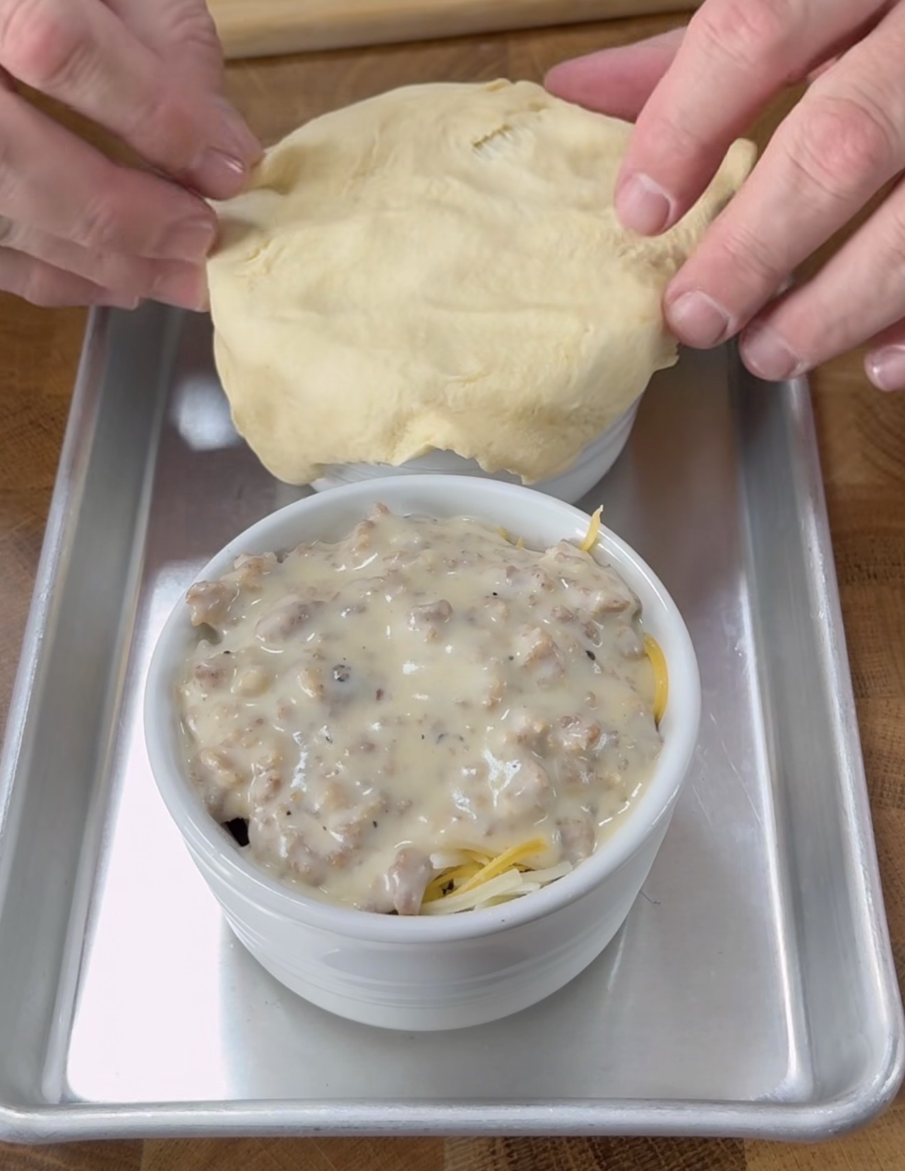 A person places a round piece of dough over a ramekin filled with sausage gravy and shredded cheese, preparing to cover it. The ramekin sits on a metal baking tray.