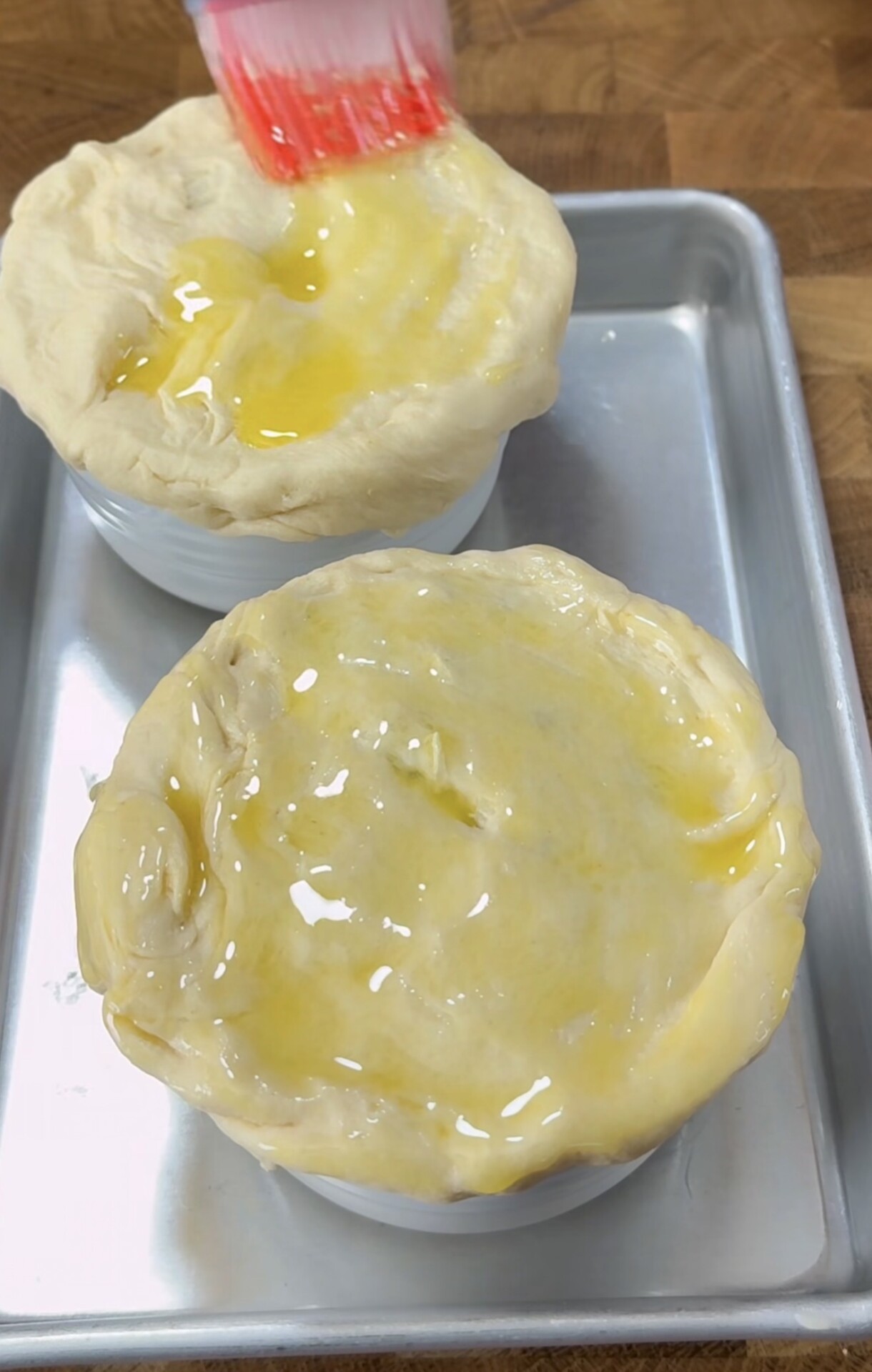 Two small ramekins are topped with dough, which is being brushed with a yellow egg wash using a red pastry brush. The ramekins sit on a metal baking sheet on a wooden surface.