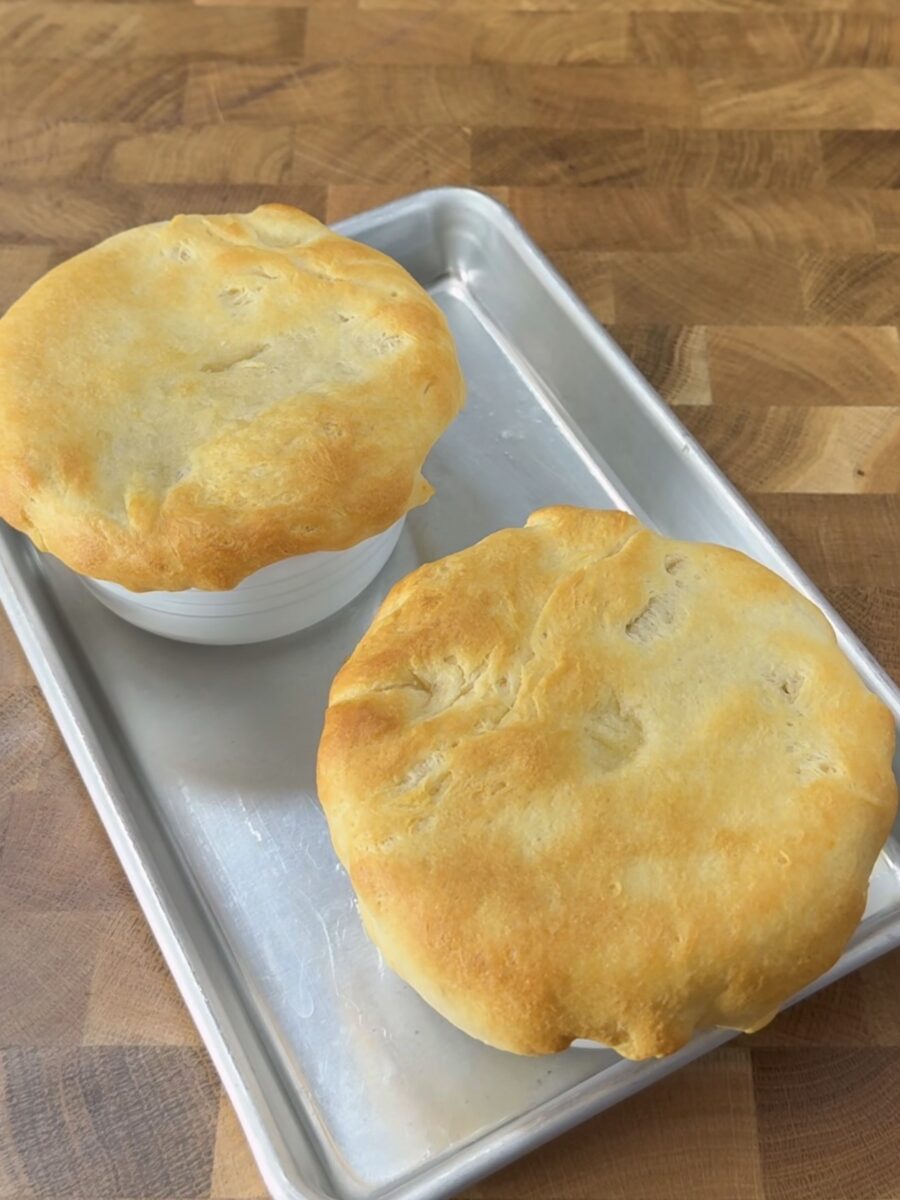 Two white ramekins filled with food, each topped with a golden-brown, baked crust, sit on a silver baking tray on a wooden surface.
