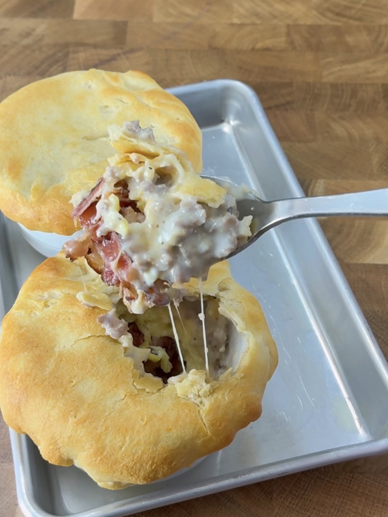 A fork lifts a portion of a creamy, cheesy pot pie with a golden, flaky crust, revealing a filling of meat and sauce inside a white ramekin on a metal tray atop a wooden surface.