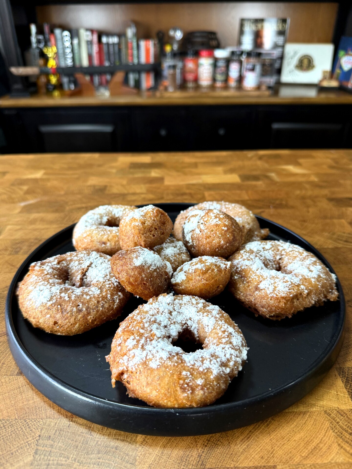 A black plate with homemade donuts and donut holes dusted with powdered sugar sits on a wooden countertop, with shelves of books and various jars blurred in the background.