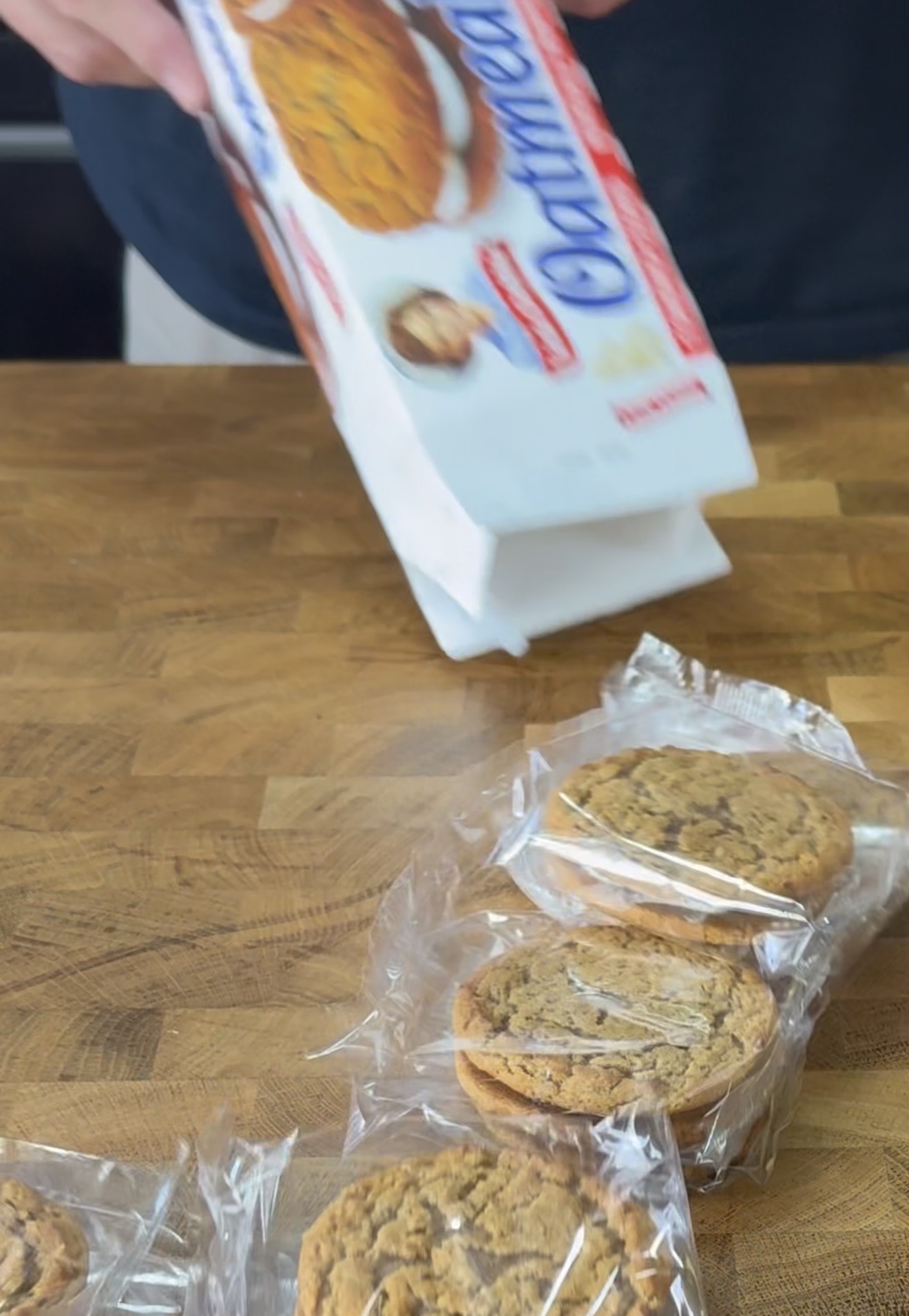 A person is opening a box of oatmeal cream pies on a wooden surface, with several individually wrapped cookie sandwiches visible outside the box.