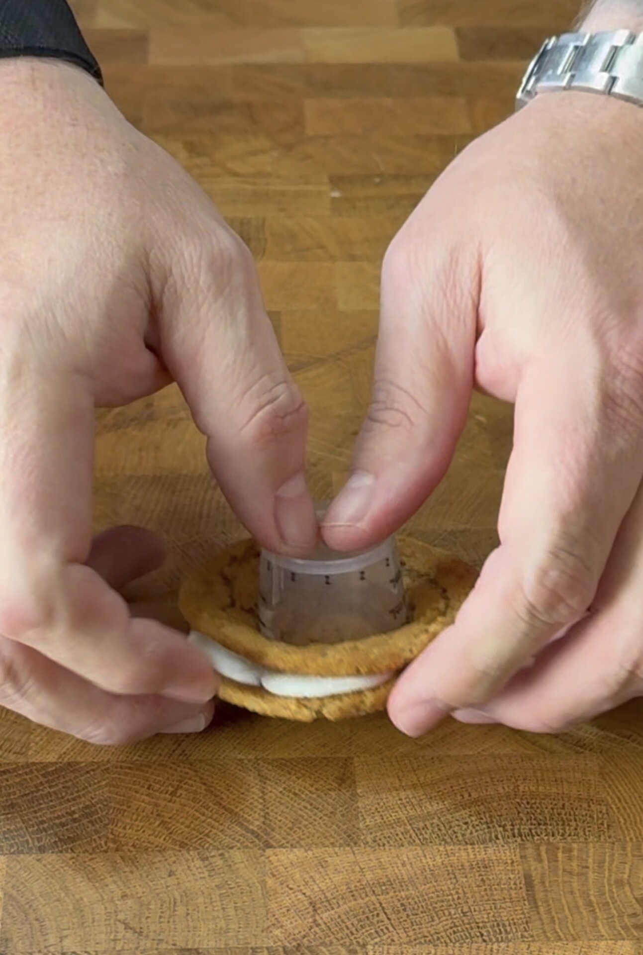 A person uses a round cookie cutter to shape a sandwich cookie with a cream filling on a wooden surface.
