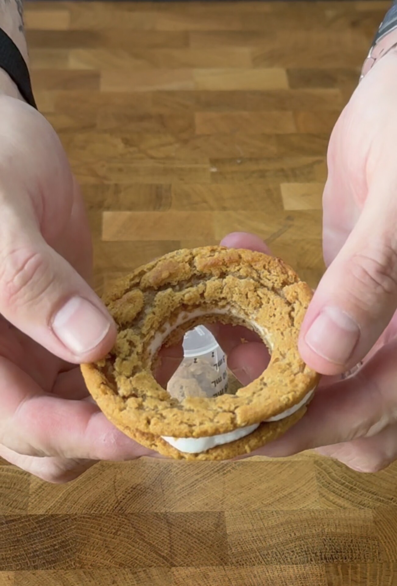 A person holds a sandwich cookie with a large circular hole cut out of the center, revealing a wooden surface below.