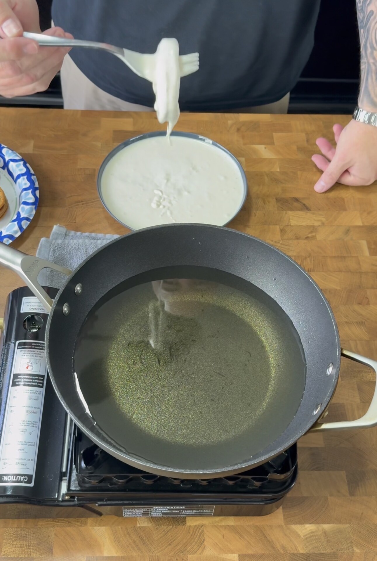 A person holds a battered food item with a fork over a bowl of batter, preparing to fry it in a pan of hot oil on a stovetop. A paper plate with food is visible on the wooden counter.