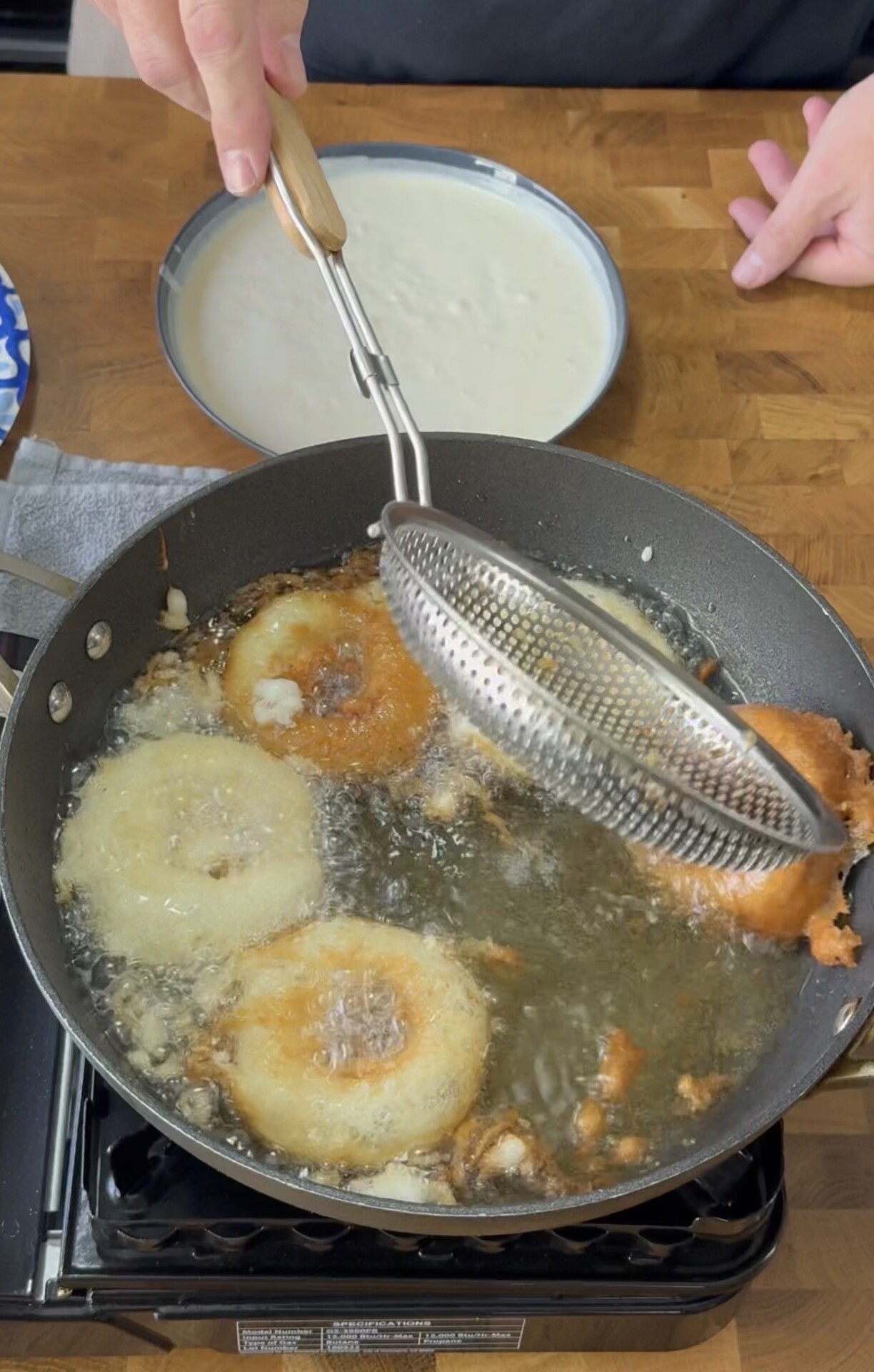 A person uses a metal skimmer to fry battered onion rings in hot oil on a stovetop. A plate with white batter and a blue-patterned dish are visible on the wooden countertop nearby.
