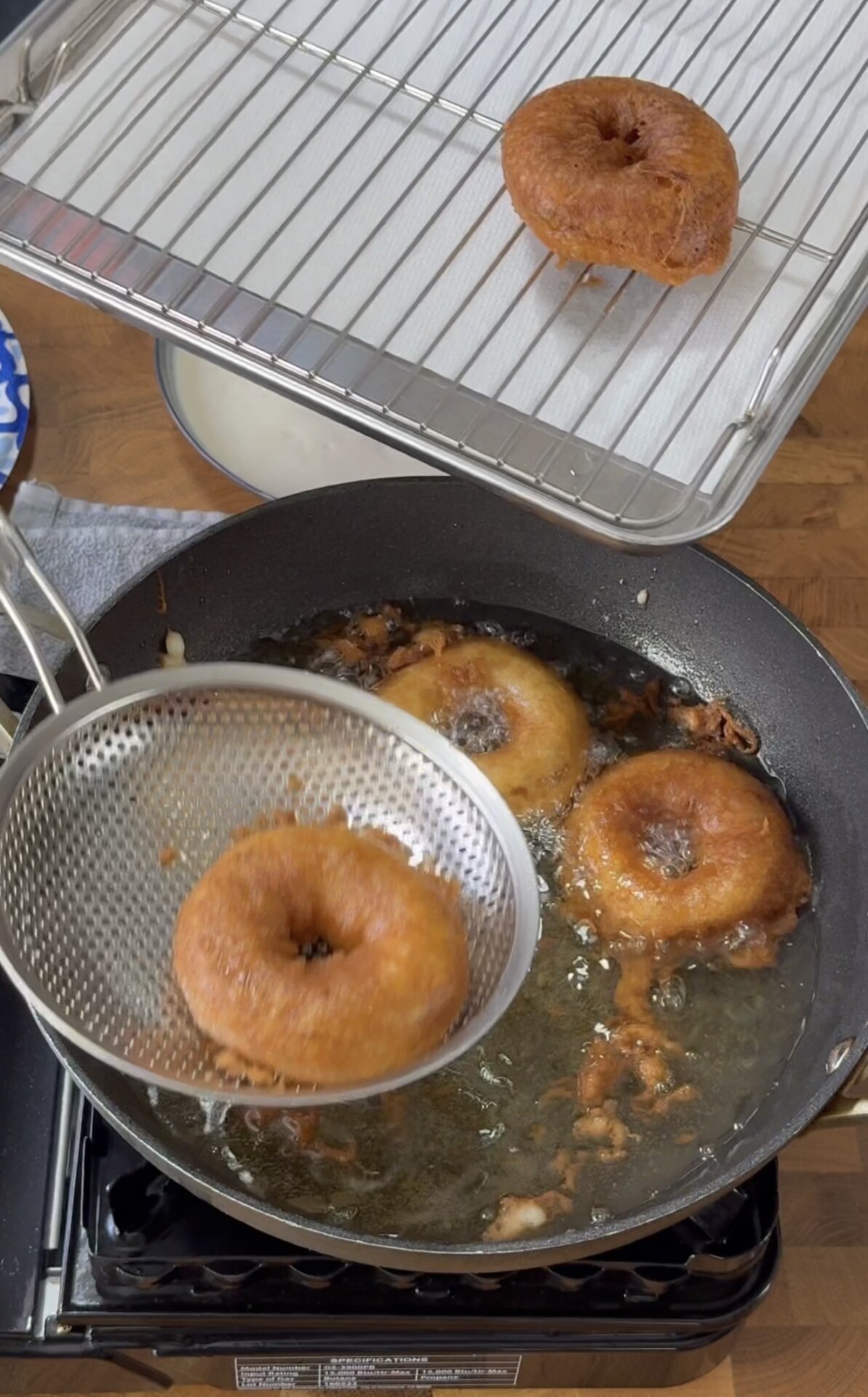 Three donuts are being deep-fried in a pan of oil, with one donut being lifted out using a slotted spoon. A cooling rack with a paper towel and a finished donut sits above the pan.