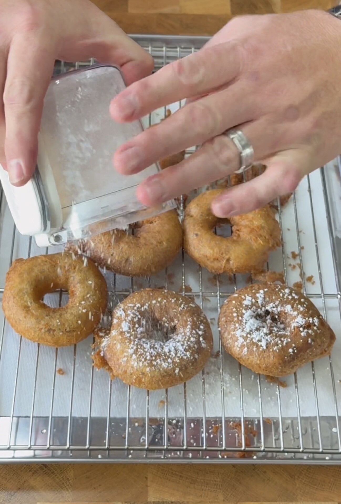 A person sprinkles powdered sugar over freshly fried doughnuts resting on a wire rack above a baking sheet.