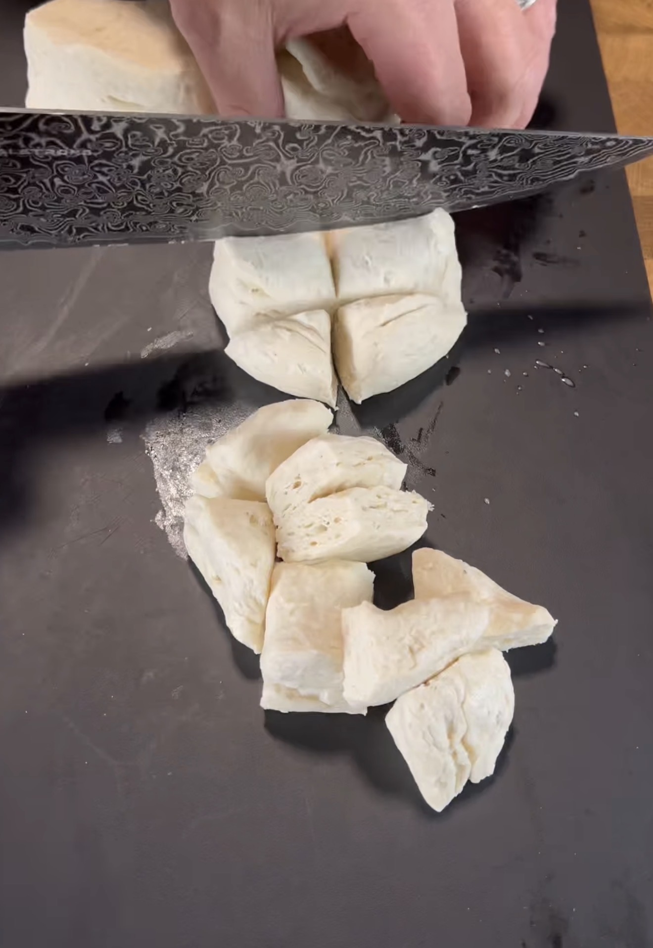 A hand uses a patterned knife to cut uncooked biscuit dough into smaller pieces on a dark cutting board.