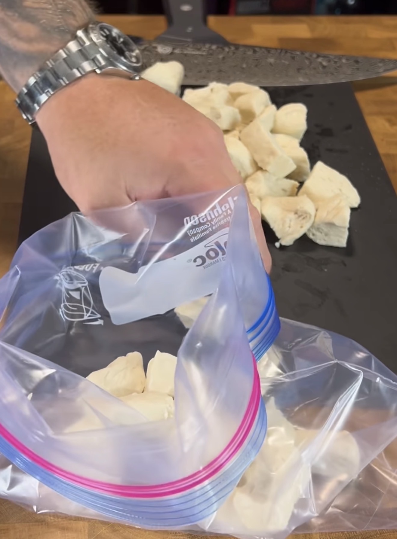 A person places pieces of chopped chicken into a resealable plastic bag, with more chicken pieces and a large knife on a black cutting board in the background.