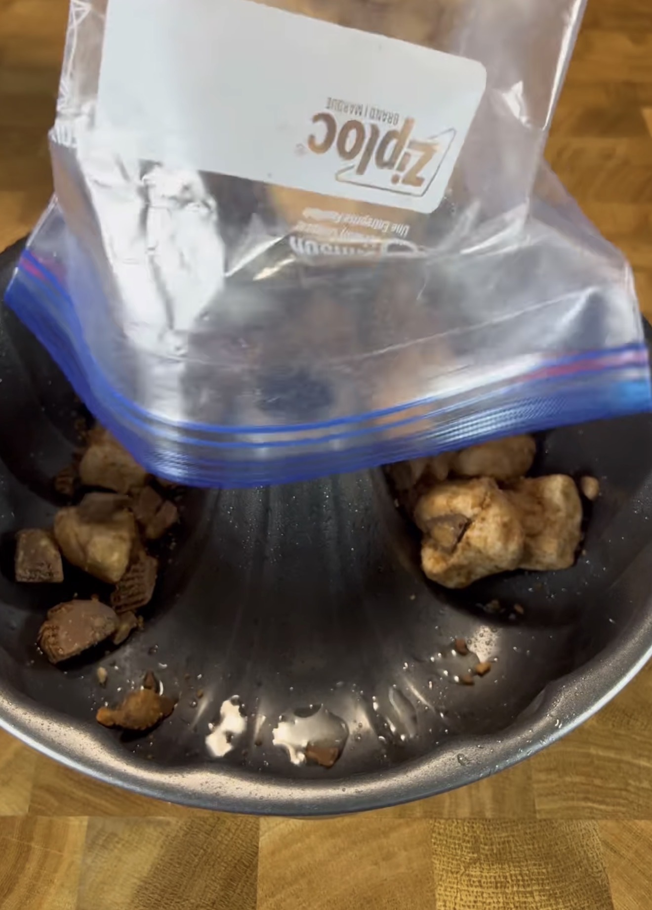 A Ziploc bag with cookies inside is being pressed into a greased Bundt pan, crushing the cookies against the pan’s surface on a wooden countertop.