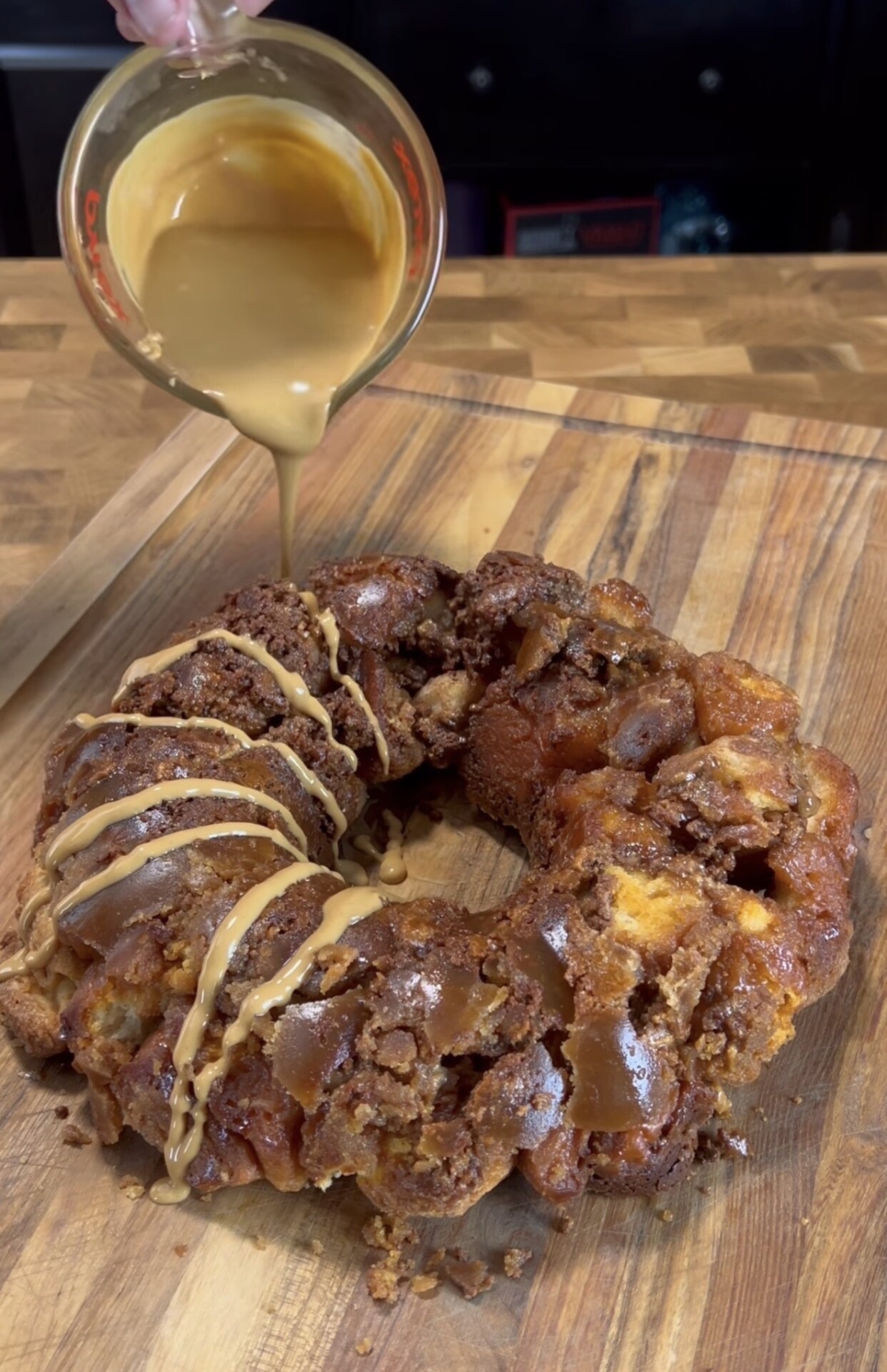A caramel sauce is being poured from a measuring cup onto a round, pull-apart monkey bread loaf on a wooden cutting board. The bread appears golden-brown with a crunchy topping.