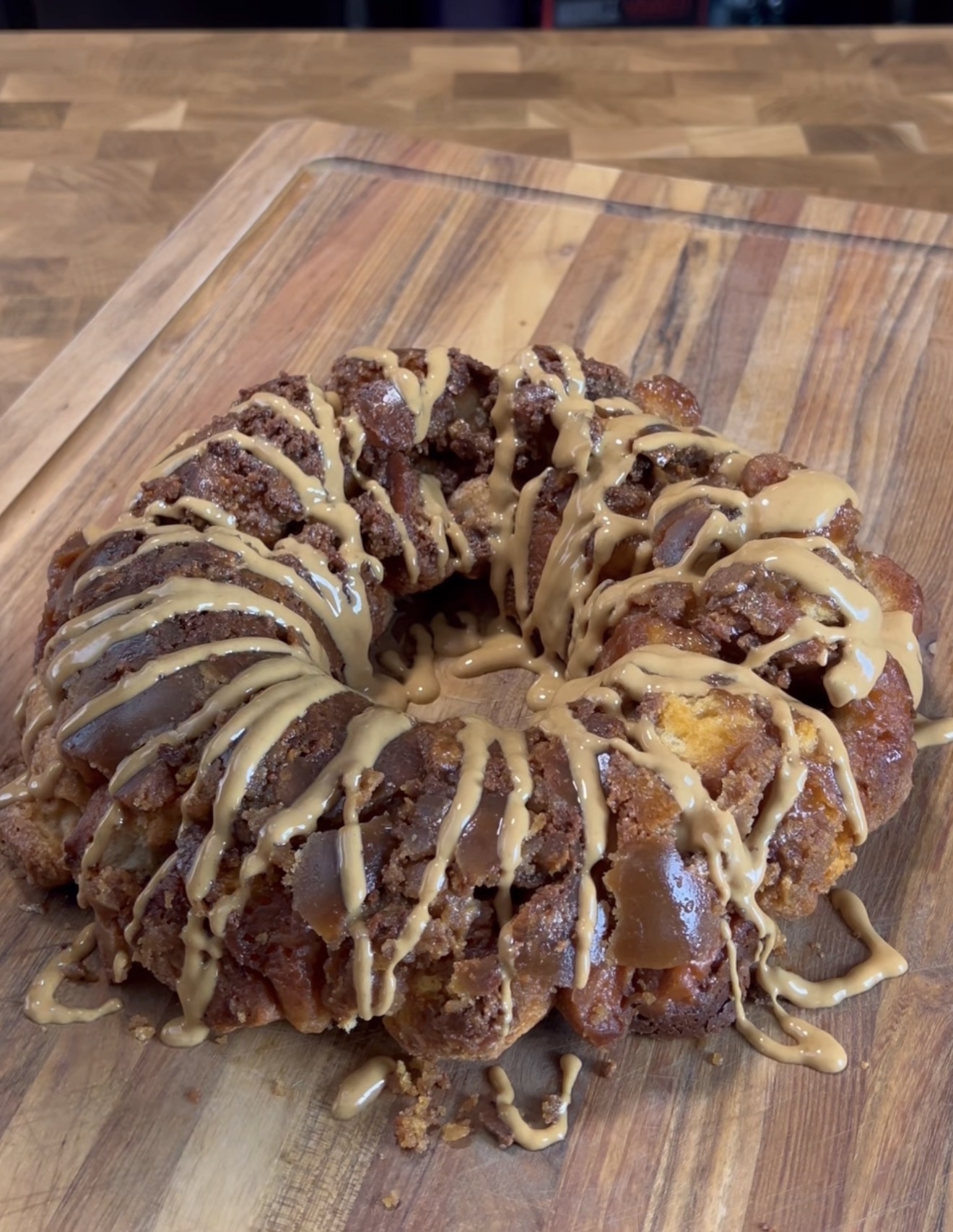 A round monkey bread loaf sits on a wooden cutting board, topped with a generous drizzle of light brown icing. The bread has a golden-brown, textured appearance with pieces baked together.