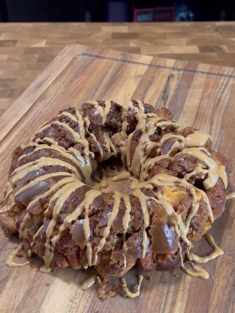 A round monkey bread dessert sits on a wooden cutting board, topped with a generous drizzle of light brown glaze. The bread pieces appear golden and caramelized.