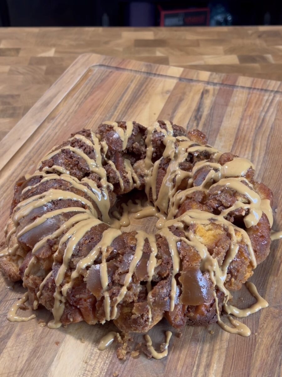 A round monkey bread dessert sits on a wooden cutting board, topped with a generous drizzle of light brown glaze. The bread pieces appear golden and caramelized.