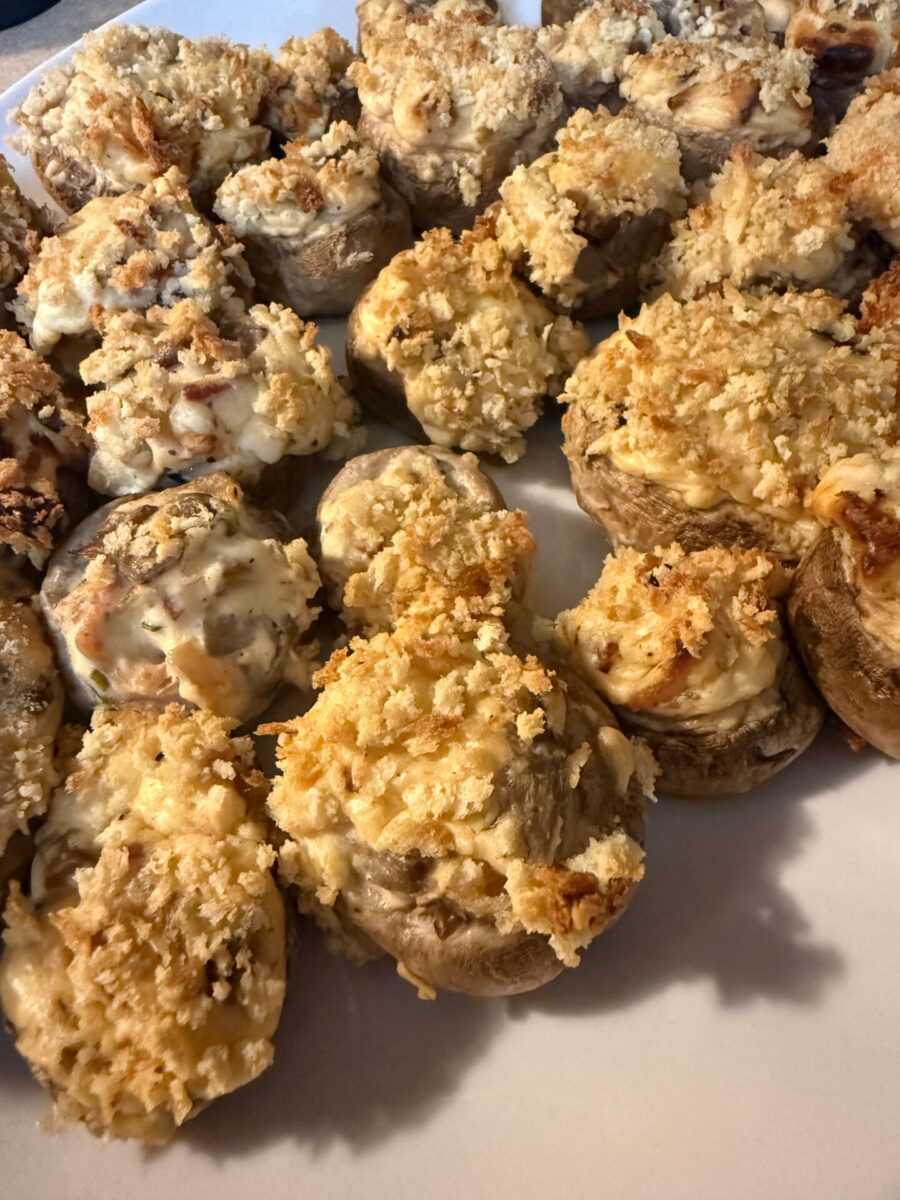 A close-up of a plate filled with baked stuffed mushrooms topped with golden-brown breadcrumbs and melted cheese. The mushrooms are arranged closely together on a white surface.