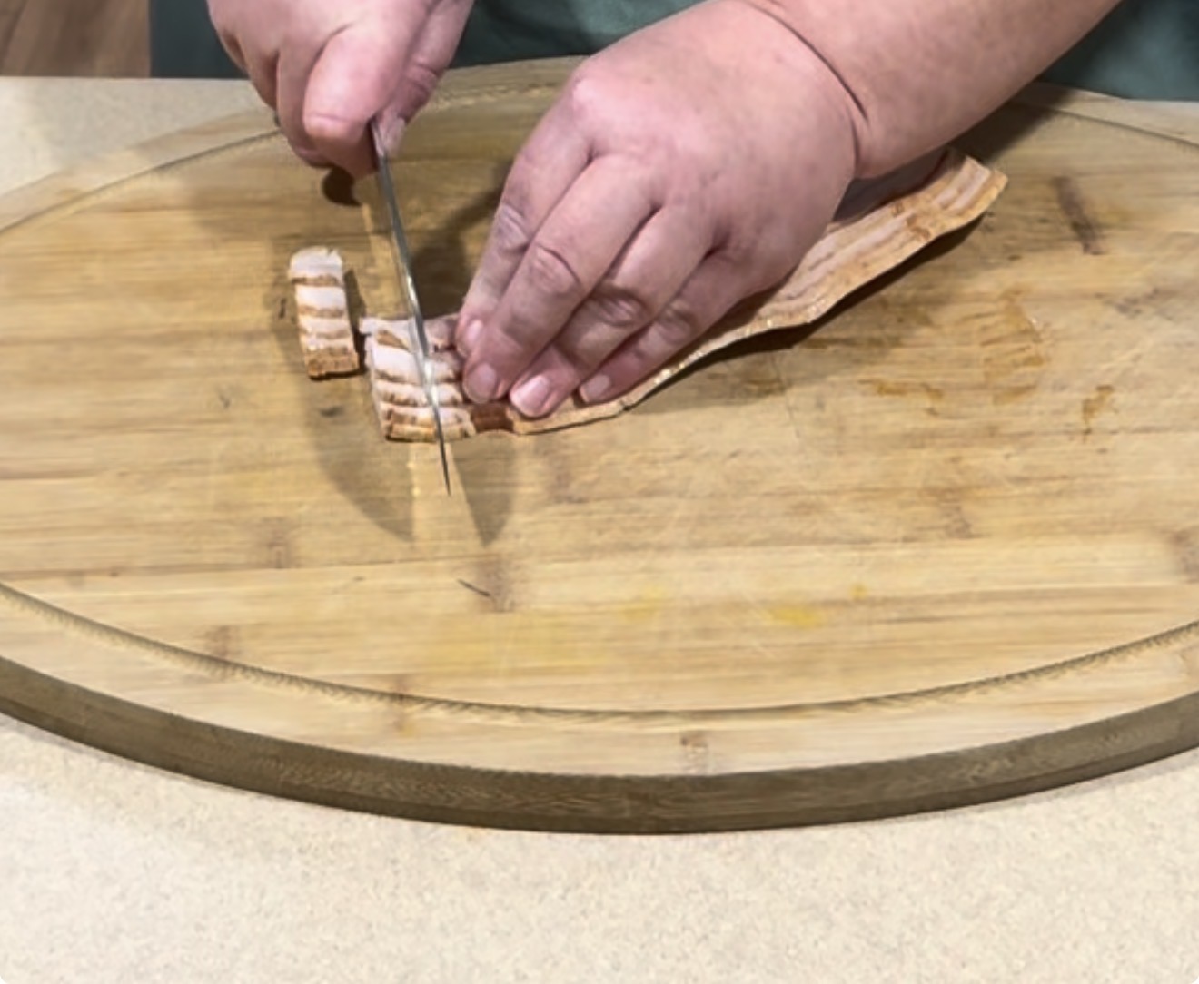 A person slices raw bacon into small pieces on a large wooden cutting board using a knife.