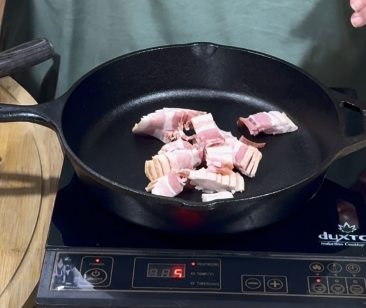Chopped raw bacon pieces in a black cast iron skillet on an induction cooktop, with the heat setting displayed as 5.
