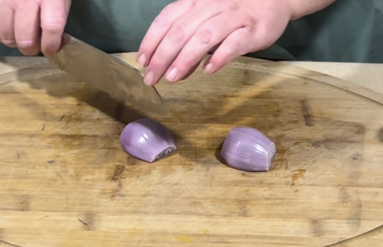 A person uses a large knife to chop a halved red onion on a wooden cutting board.