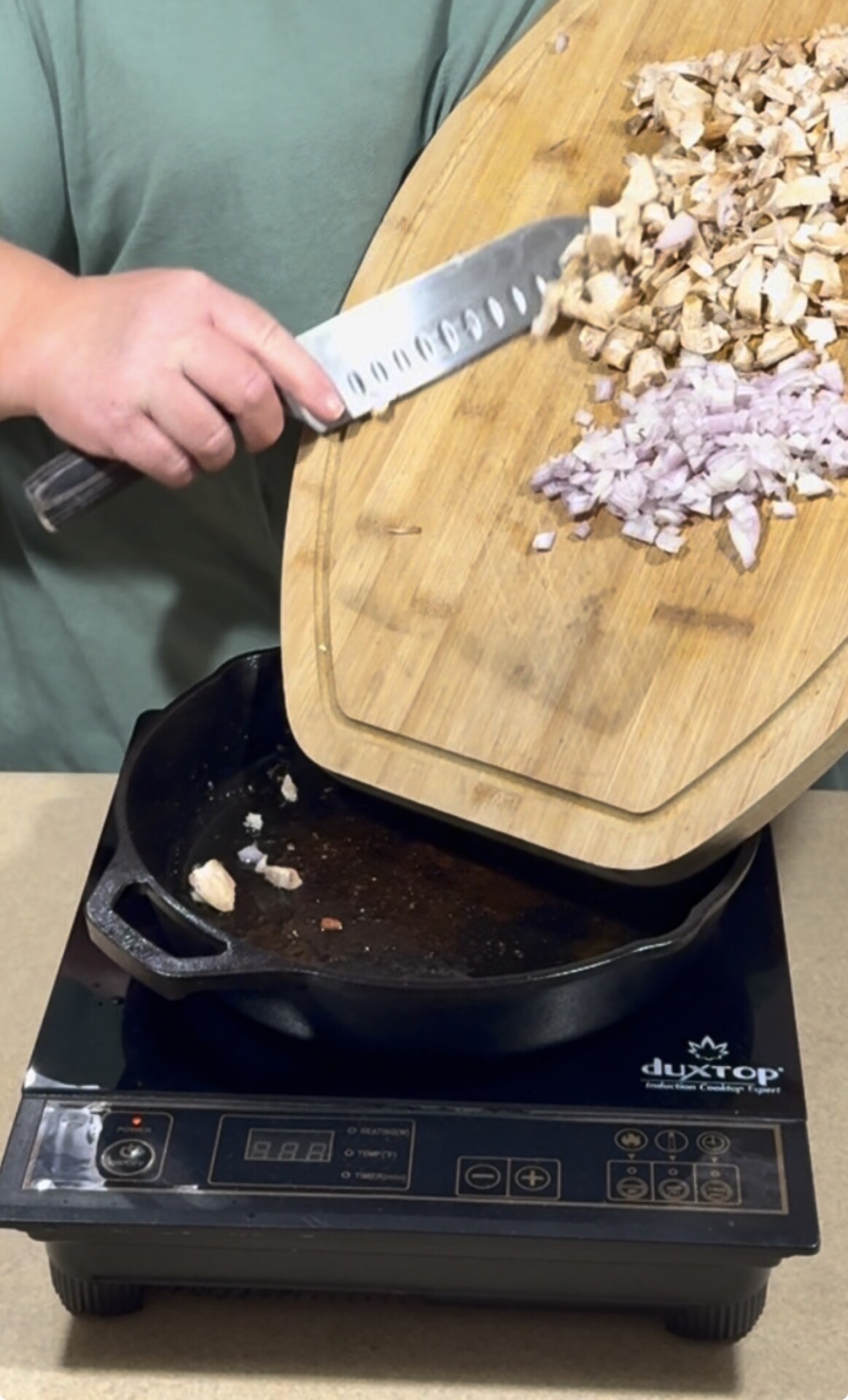 A person slides chopped mushrooms and onions from a wooden cutting board into a black skillet on an induction cooktop.