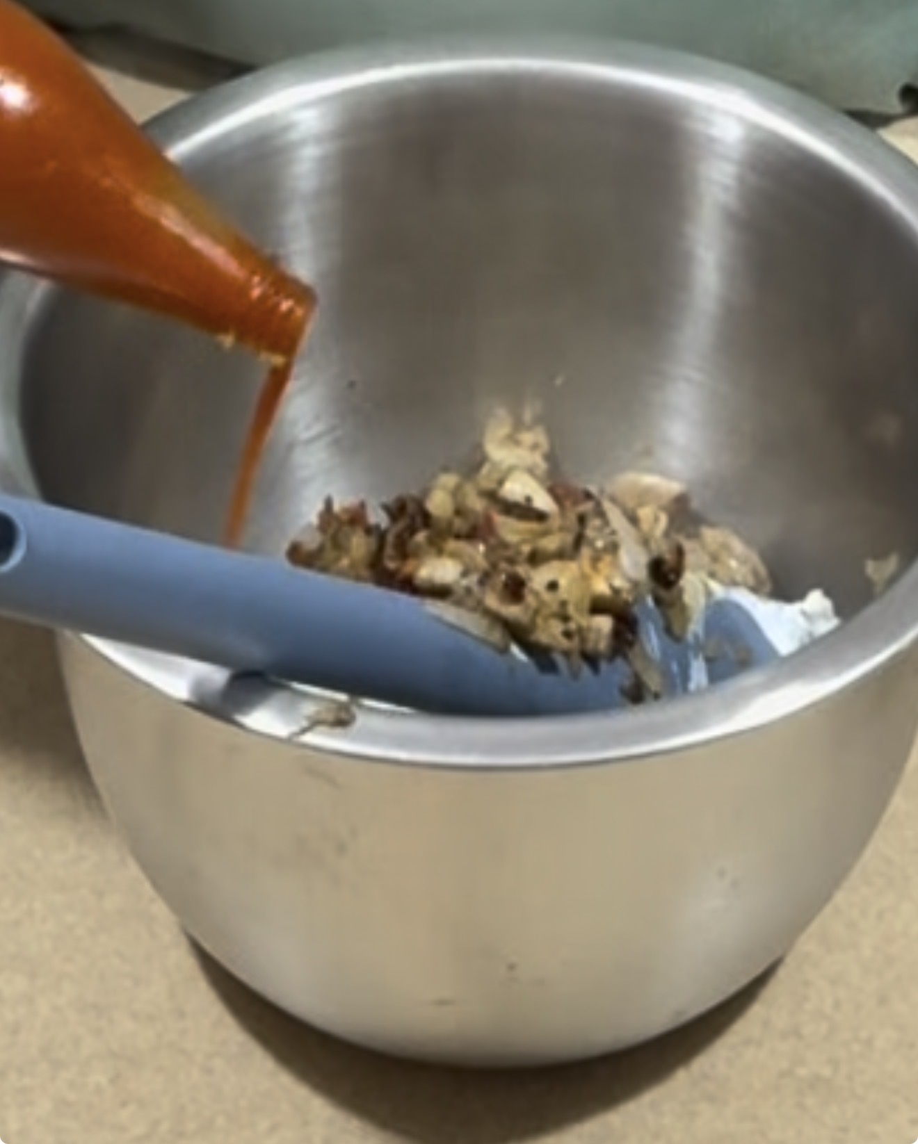 A bottle of hot sauce is being poured into a metal mixing bowl containing a mixture of chopped vegetables or mushrooms, with a blue spatula resting inside the bowl.