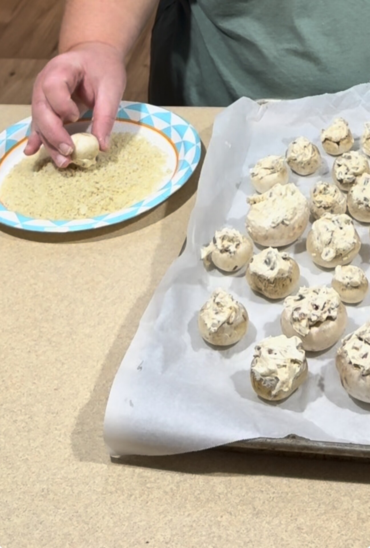A person dips a stuffed mushroom into breadcrumbs on a paper plate, while a baking sheet lined with parchment paper holds several stuffed mushrooms ready to bake.