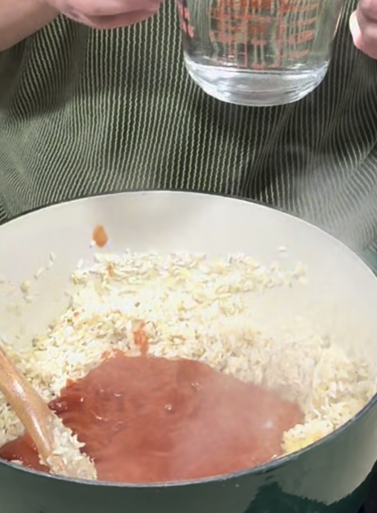 A person holds a glass measuring cup above a pot filled with rice and tomato sauce, with steam rising and a wooden spoon resting inside the pot.