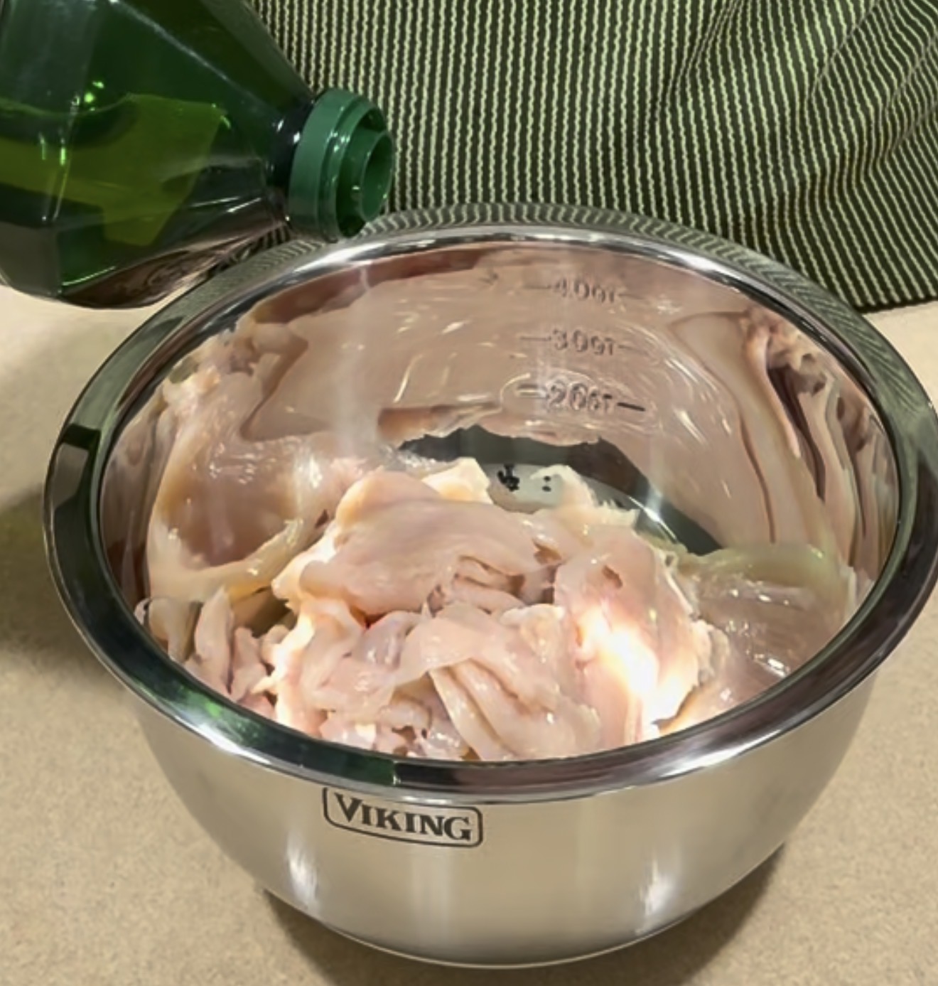 A metal bowl labeled Viking contains raw sliced chicken. Olive oil is being poured from a green bottle into the bowl. A person in a green striped shirt stands in the background.