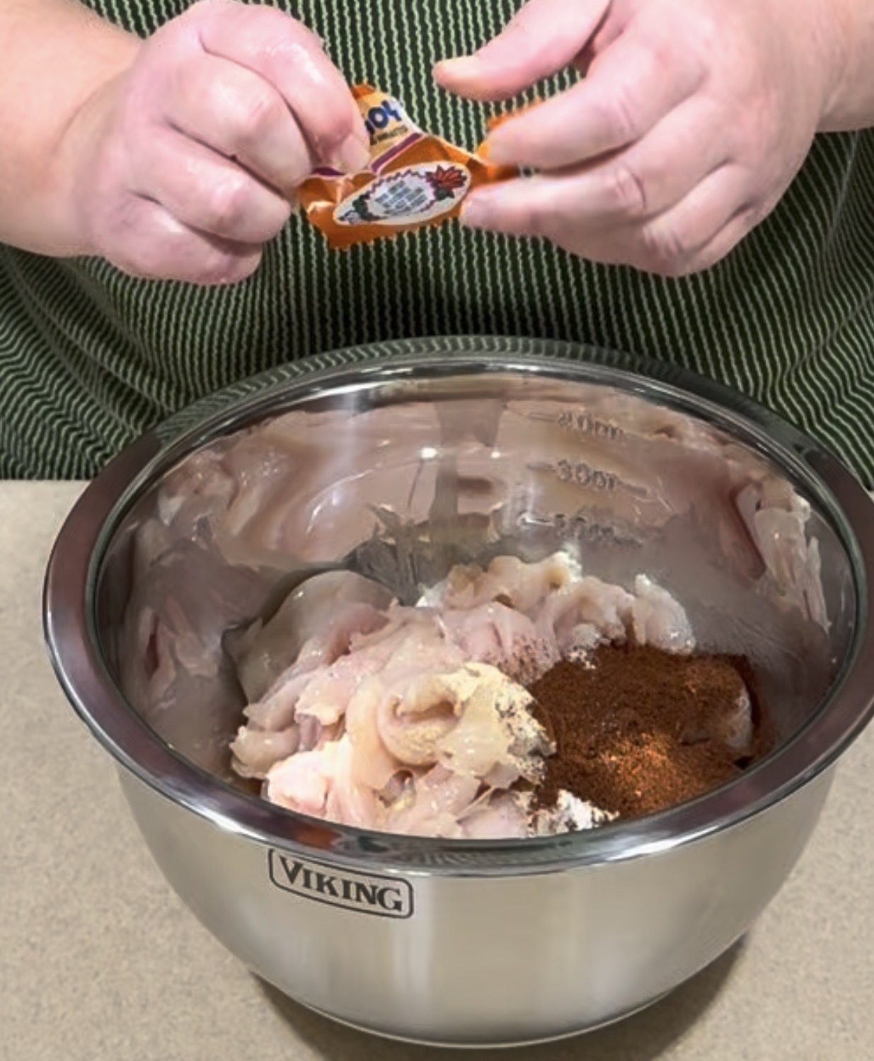 A person adds spices from a packet to a stainless steel bowl containing raw, sliced chicken and various seasonings on a kitchen counter.