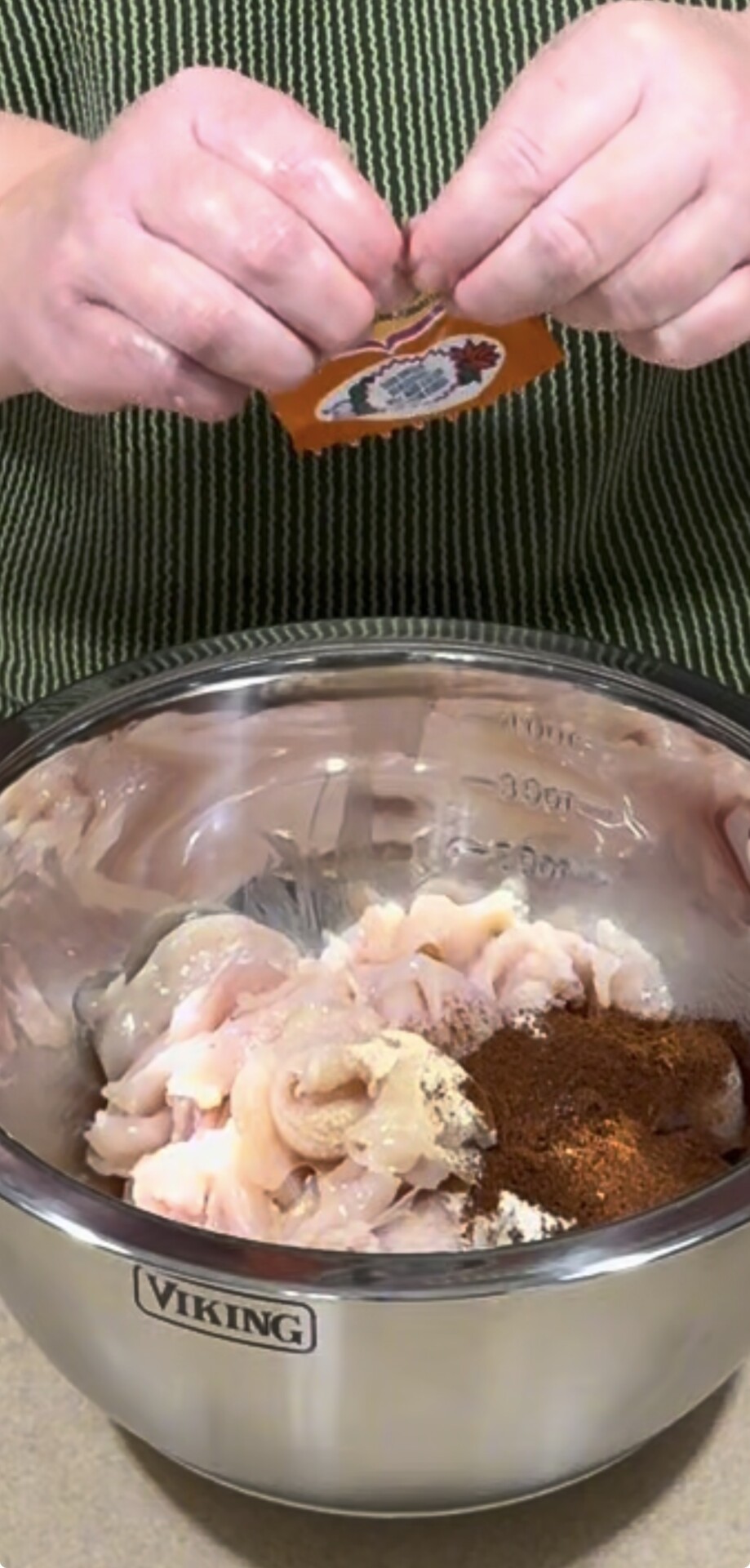 A person sprinkles spices from a small packet into a metal bowl containing raw chicken pieces and assorted seasonings, preparing to marinate the meat.