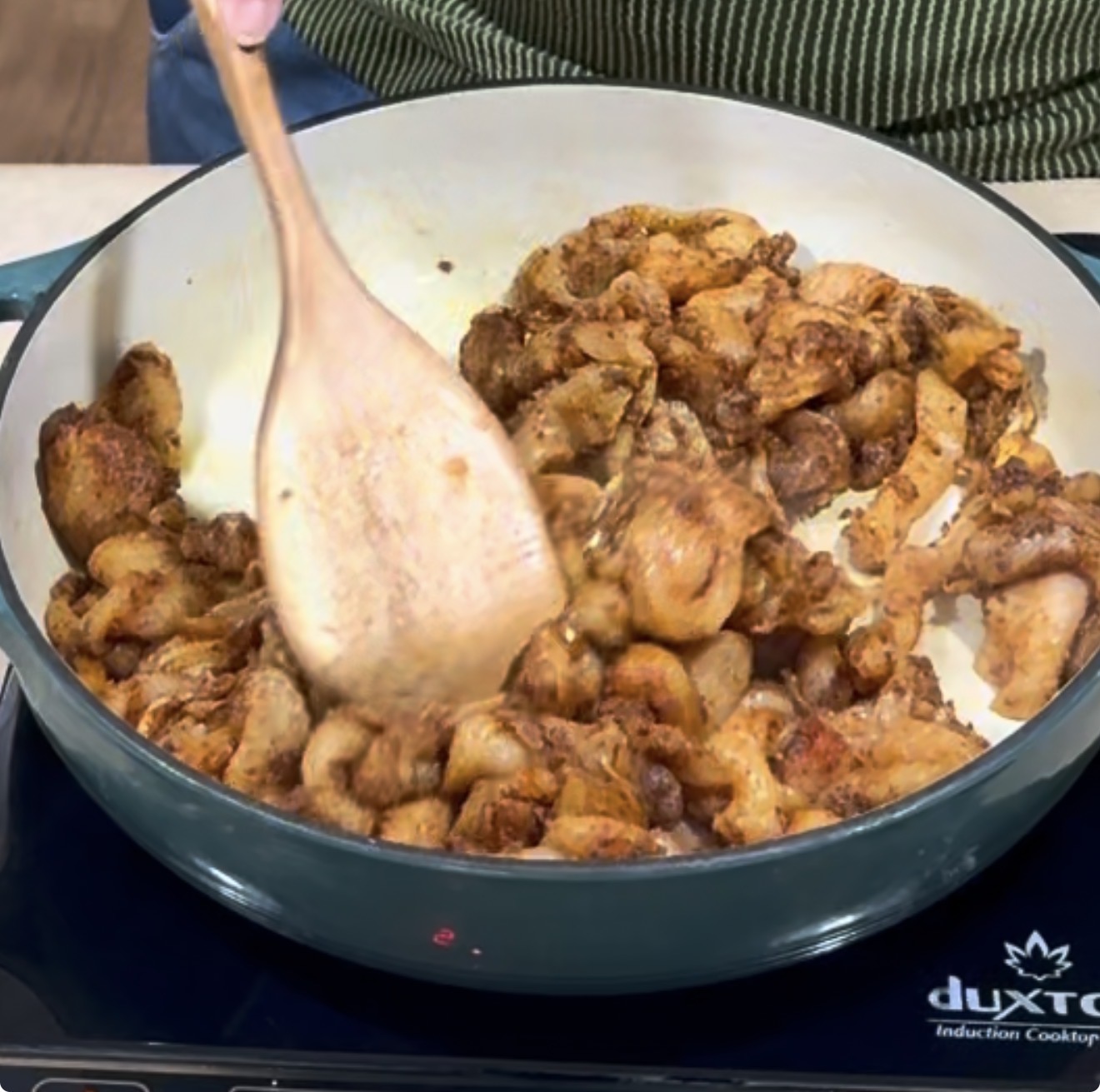 A person stirs seasoned, cooked chicken pieces in a large skillet with a wooden spoon on a Duxtop induction cooktop.