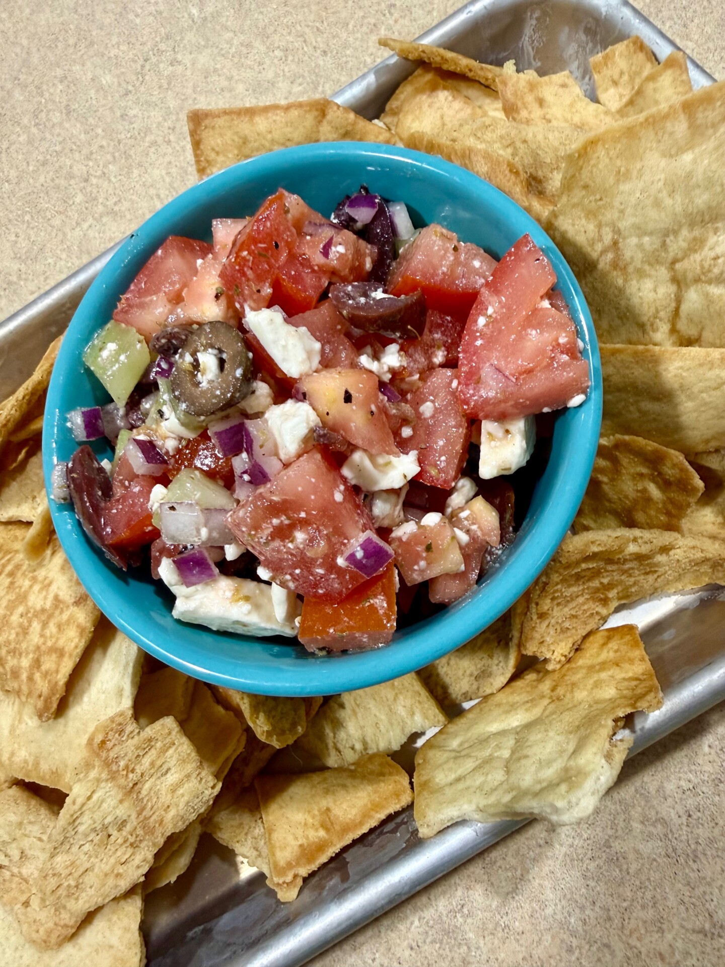 A blue bowl filled with Greek salad—featuring tomatoes, red onion, olives, cucumber, and feta—sits on a tray surrounded by crispy pita chips.